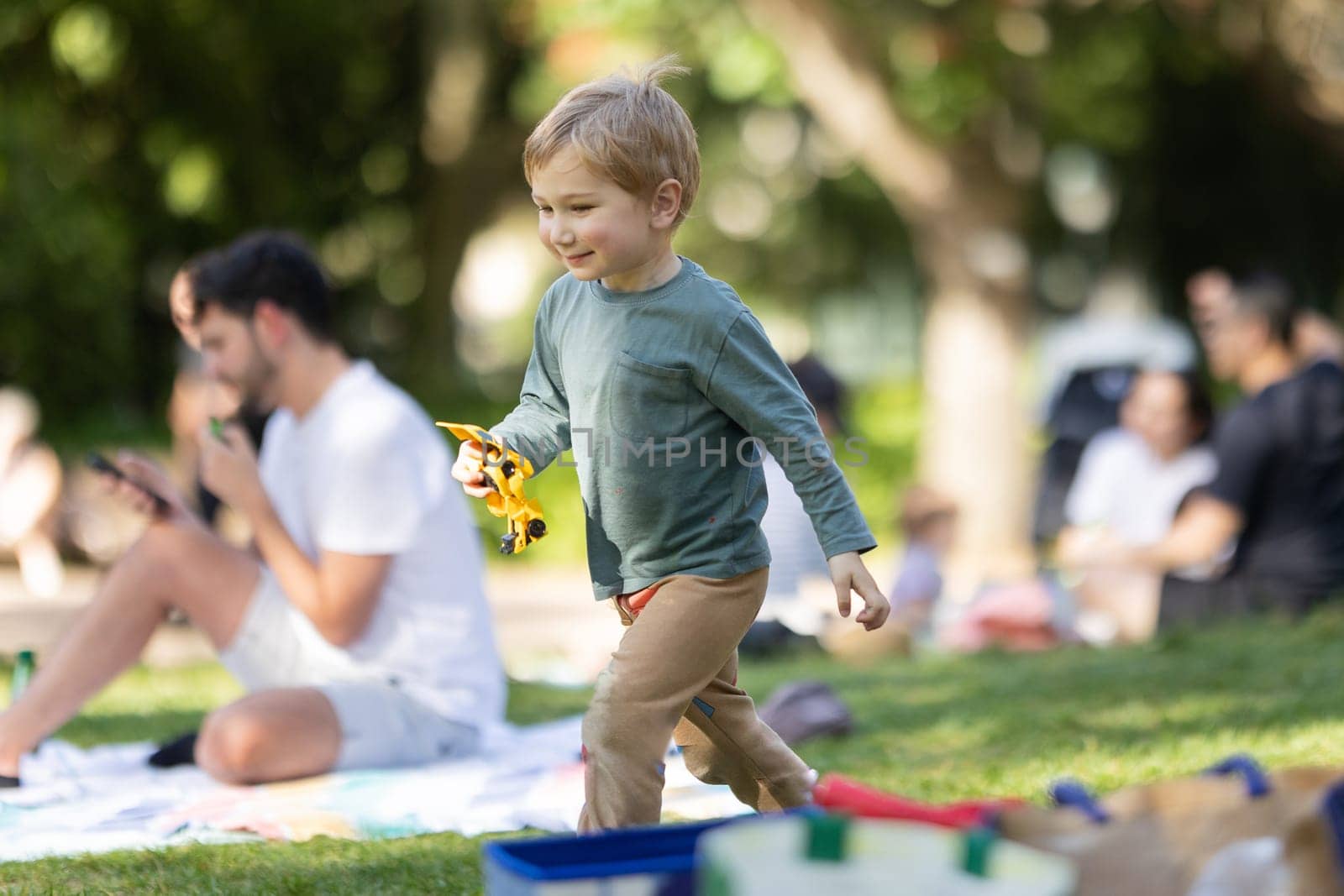 A young boy is playing with a toy truck in a park. There are other people in the background, some of whom are sitting on blankets