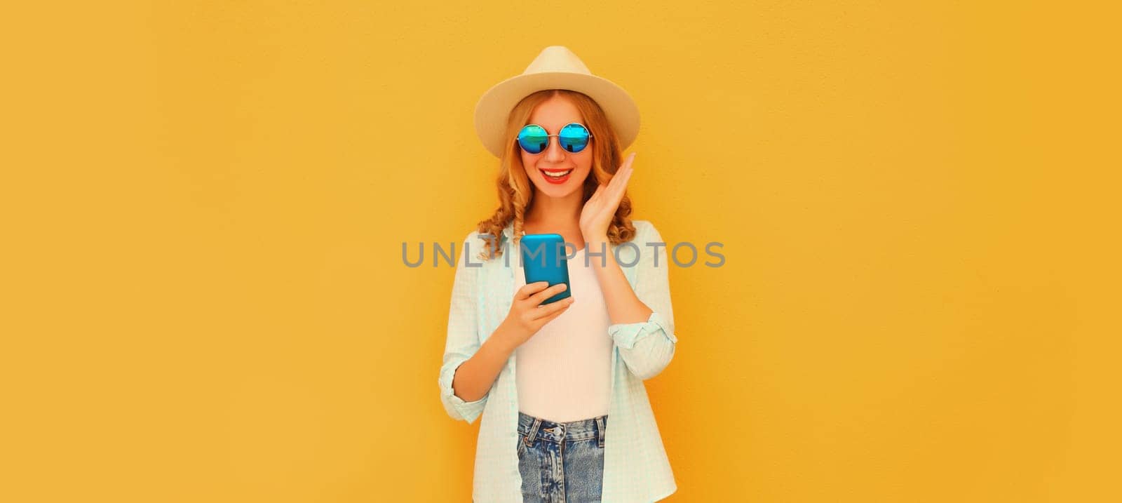 Summer portrait of happy surprised amazed young woman with mobile phone looking at device in straw tourist hat on yellow studio background