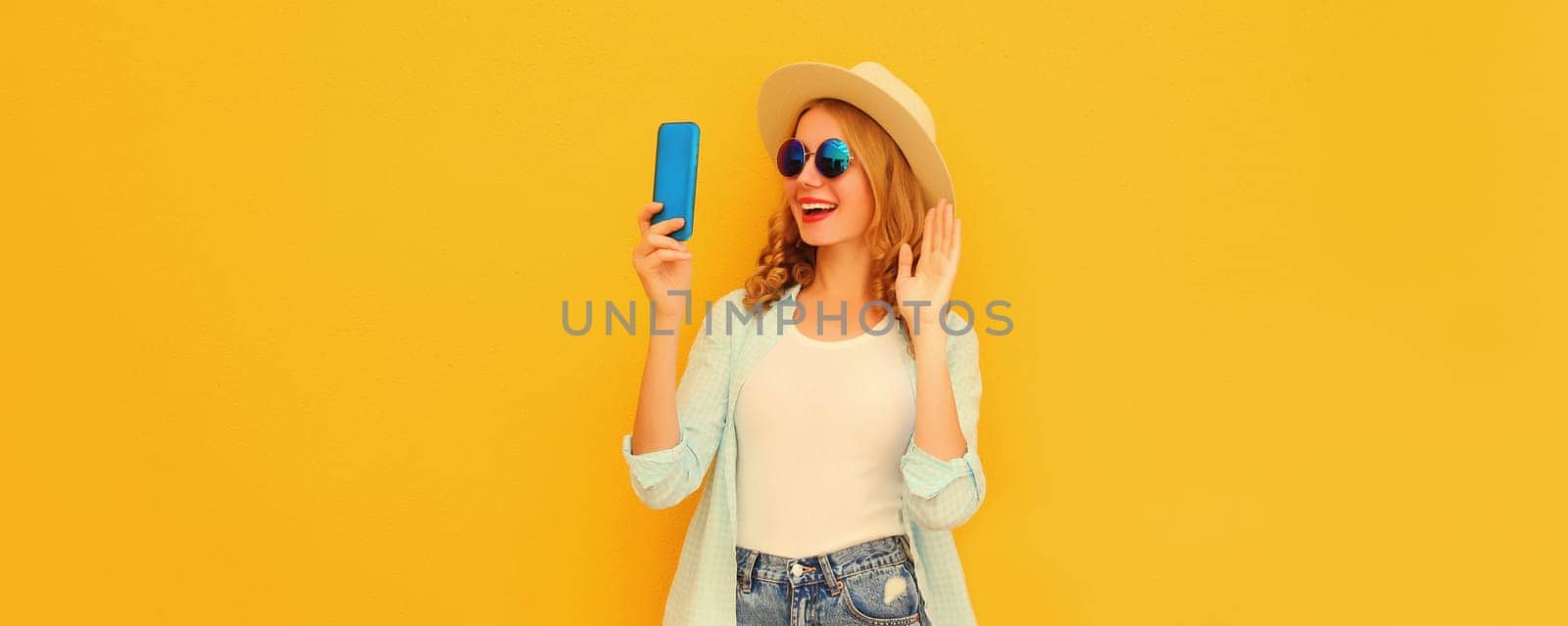 Portrait of happy joyful laughing young woman taking selfie with smartphone or talking on video call waving hand wearing summer straw hat on bright yellow background