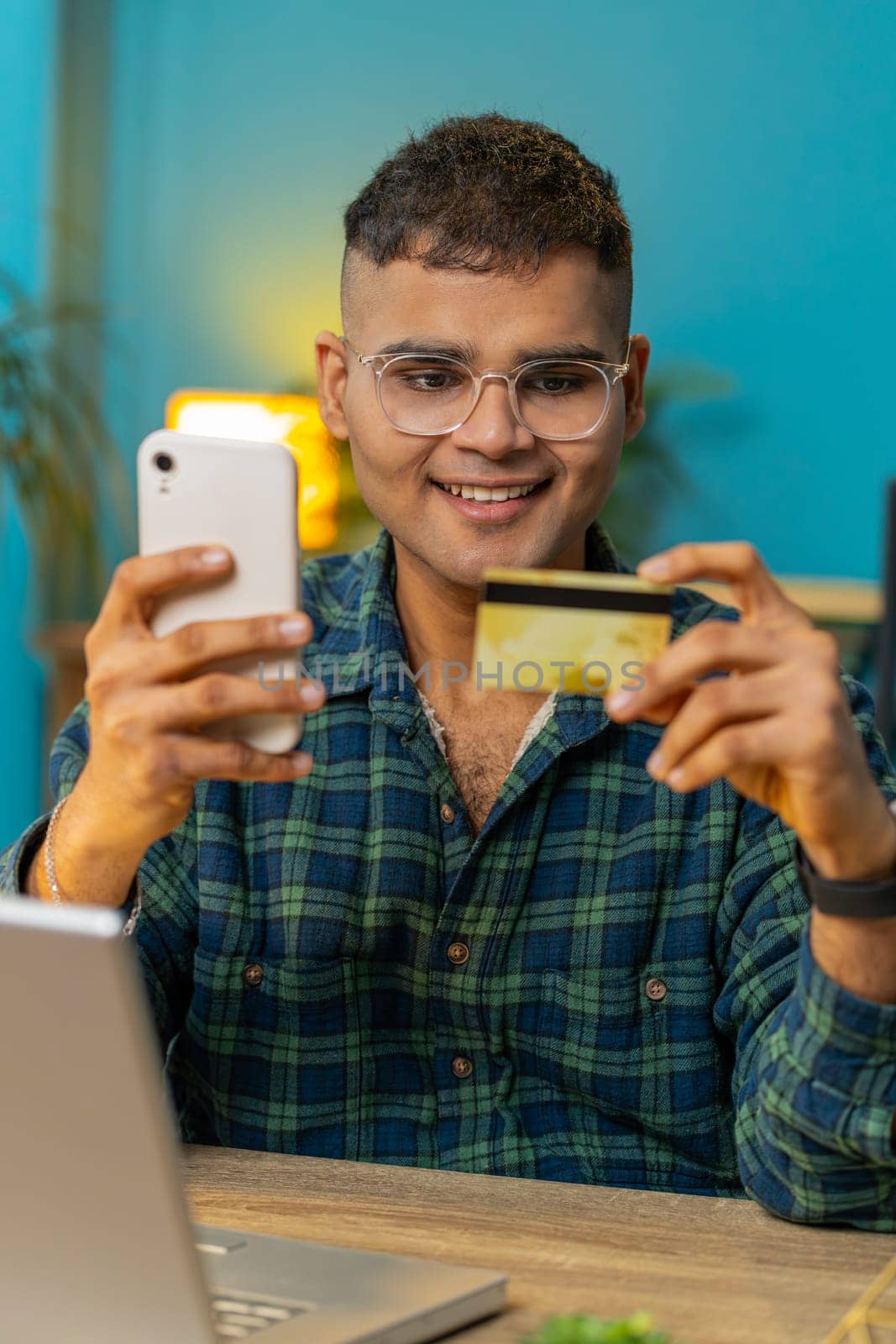 Happy Hindu man using bank credit card smartphone for online shopping payments at home office. Business transaction. Smiling guy freelancer purchases with cellphone E-banking app service. Vertical