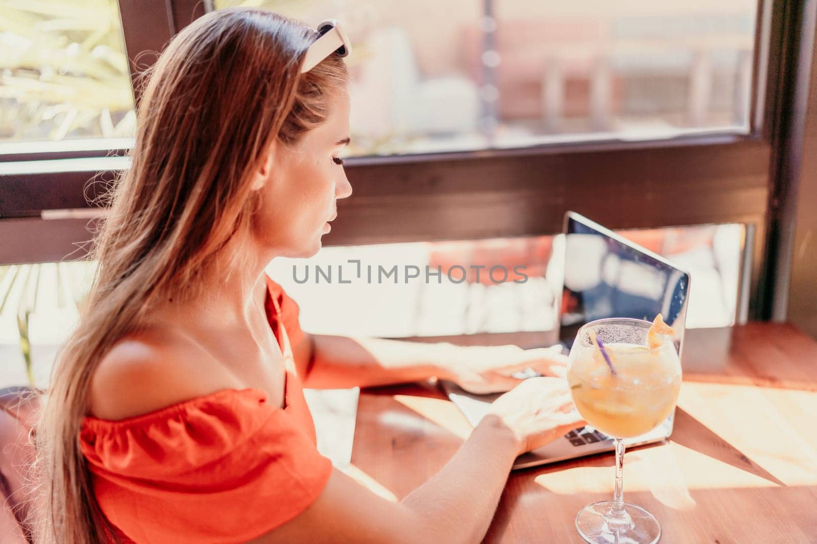 Woman works on laptop in a sunny cafe, glass of juice nearby. The setting is modern, with natural light illuminating the space. The image captures a moment of focused work