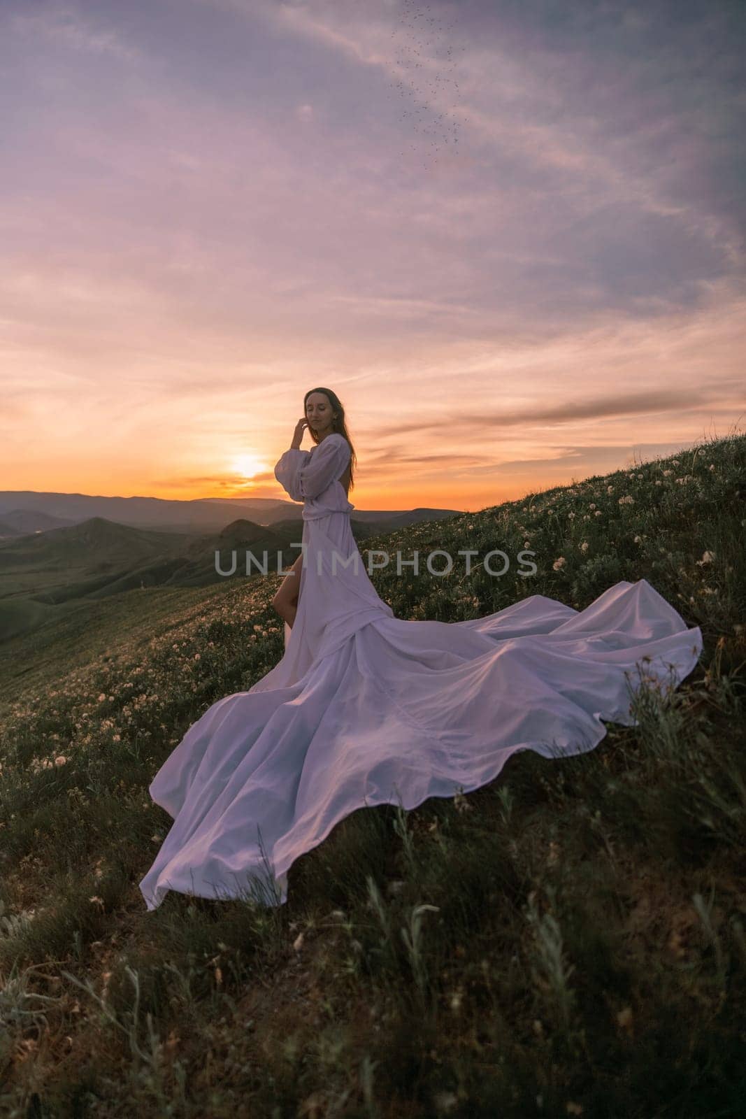 A woman in a long white dress is standing on a hillside, with the sun setting in the background. The scene is serene and peaceful, with the woman's dress flowing in the wind