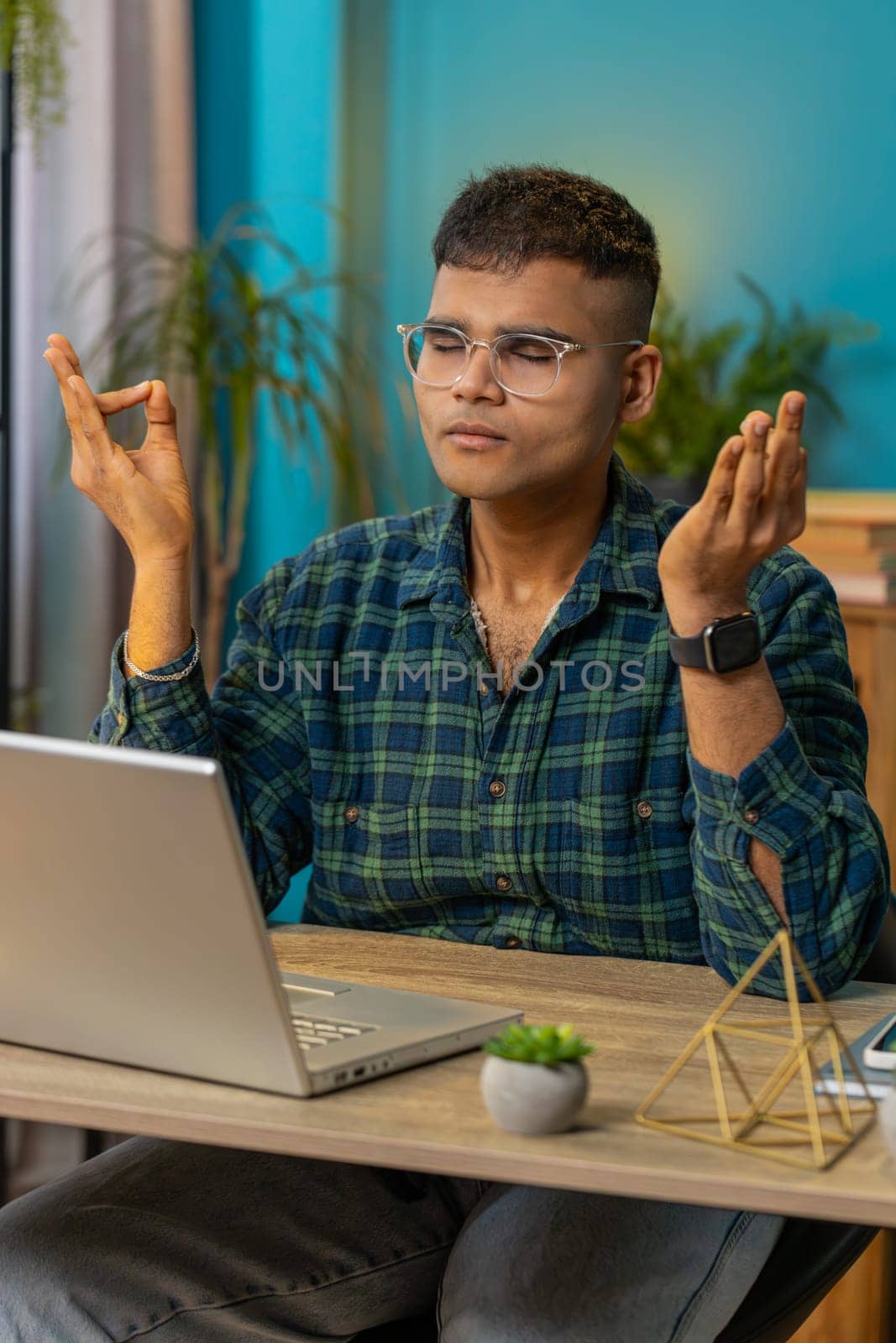 Indian man working on laptop, breathes deeply, eyes closed meditating with concentrated thoughts, peaceful mind. Hindu guy taking a break, relaxing sitting at home office table workplace. Vertical