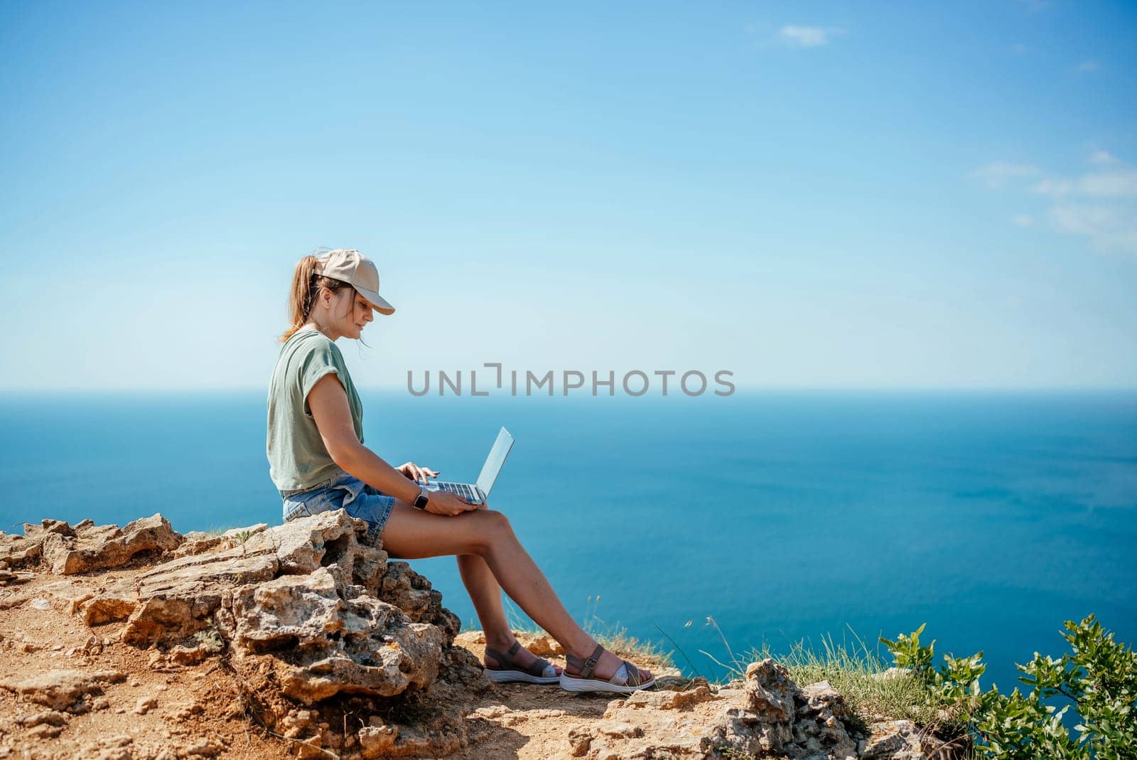 A woman is sitting on a rock overlooking the ocean with a laptop in front of her. She is working or studying, as she is focused on her laptop. Concept of solitude and tranquility