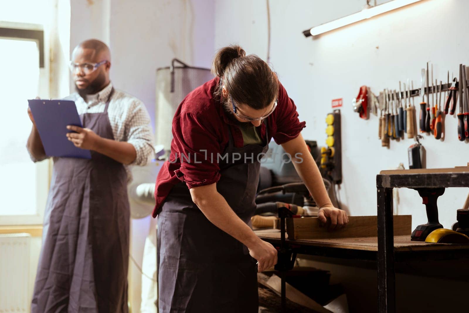 Woodworker using bench vise to hold lumber block, starting furniture assembling in workshop next to BIPOC coworker. Craftsperson using vice tool to clamp piece of wood with african american colleague