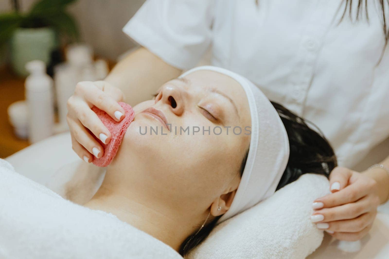 A young unrecognizable Caucasian girl cosmetologist cleans the chin of an adult female client's face using a pink scrubbing sponge, who lies with her eyes closed on a massage table in a beauty salon, side view close-up.