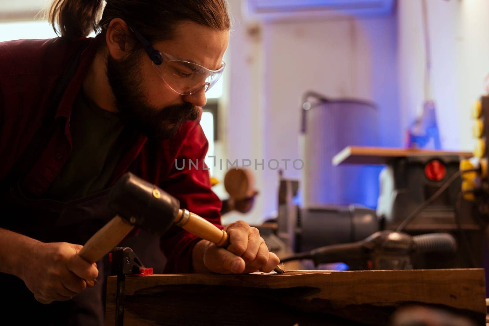 Man shaping raw timber using chisel and hammer in carpentry shop, wearing safety glasses to avoid injury. Woodworking expert making wood sculptures using protective gear to prevent workplace accidents