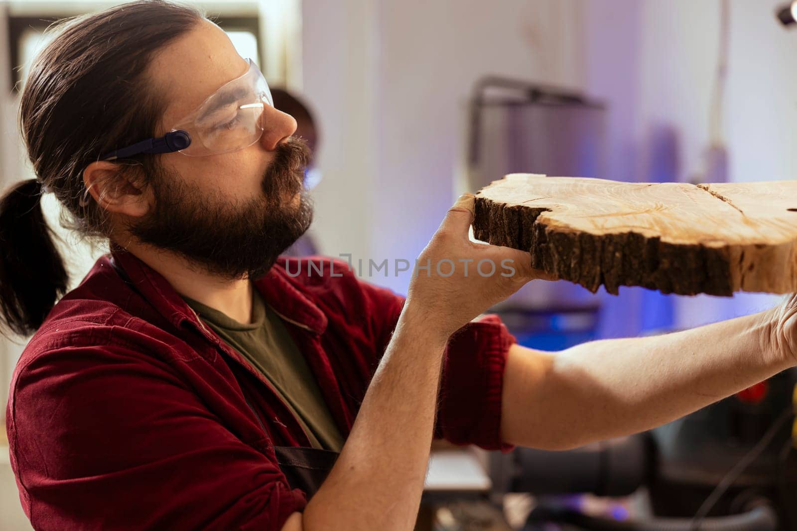 Carpenter in woodworking shop wearing protection glasses, inspecting wood before assembling furniture. Cabinetmaker using safety equipment in joinery, evaluating lumber block