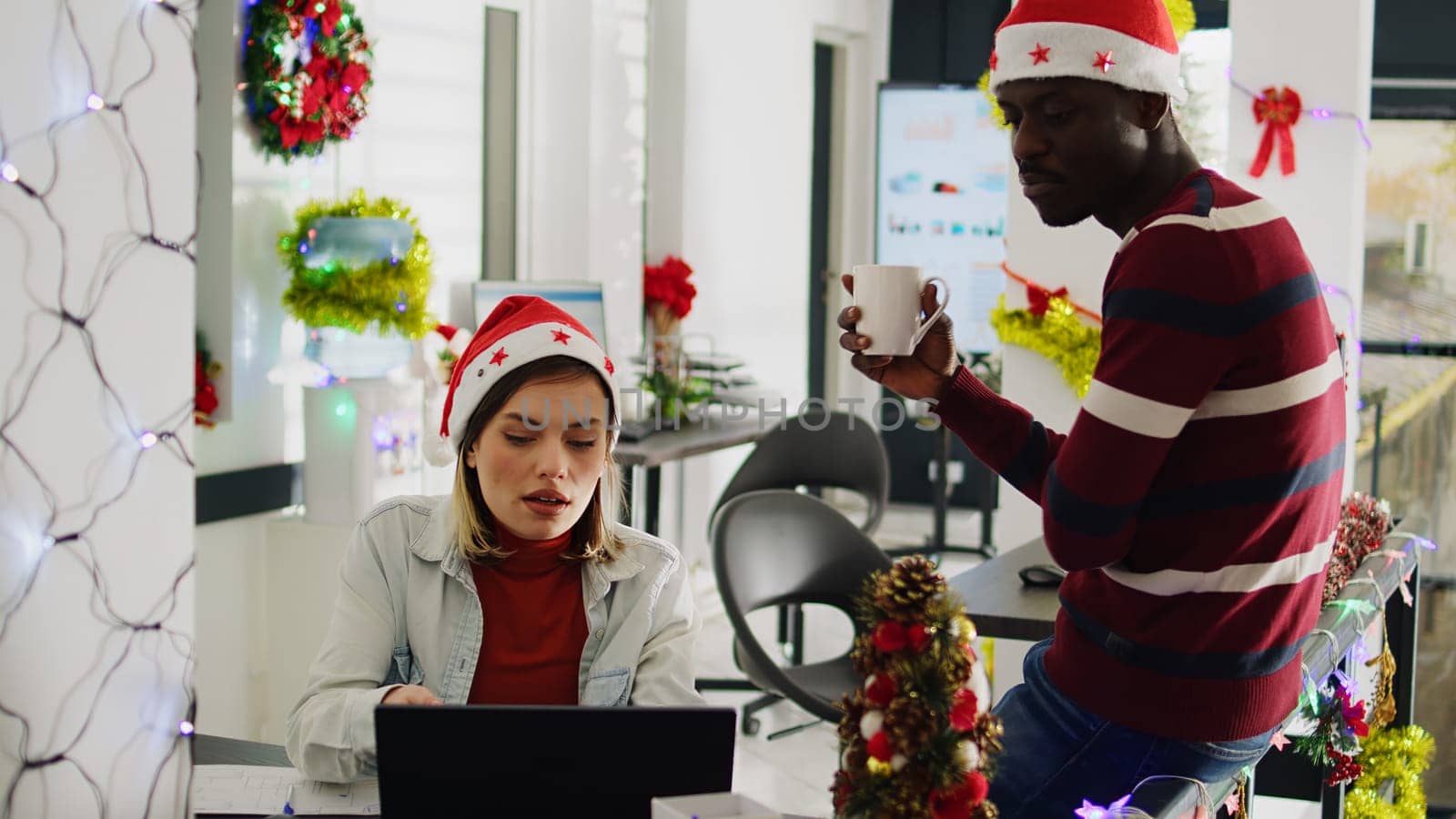 Zoom in shot of african american employee taking a break, enjoying coffee in Christmas decorated office. Staff member discussing with colleague wearing Santa hat during festive holiday season