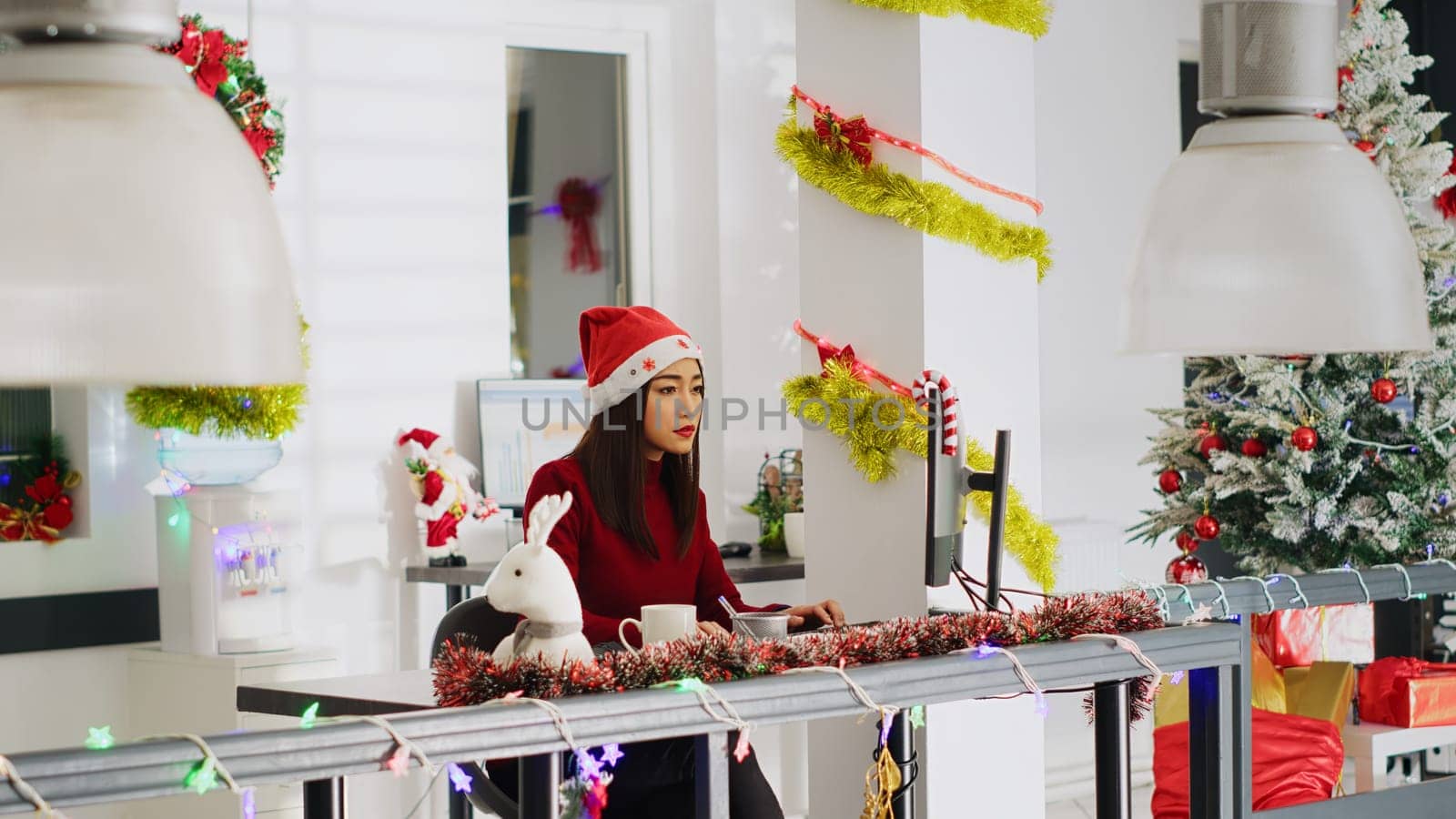 Proficient asian employee wearing Santa hat working in Christmas ornate office. Adept worker looking over business revenue numbers at computer desk in diverse xmas ornate workplace
