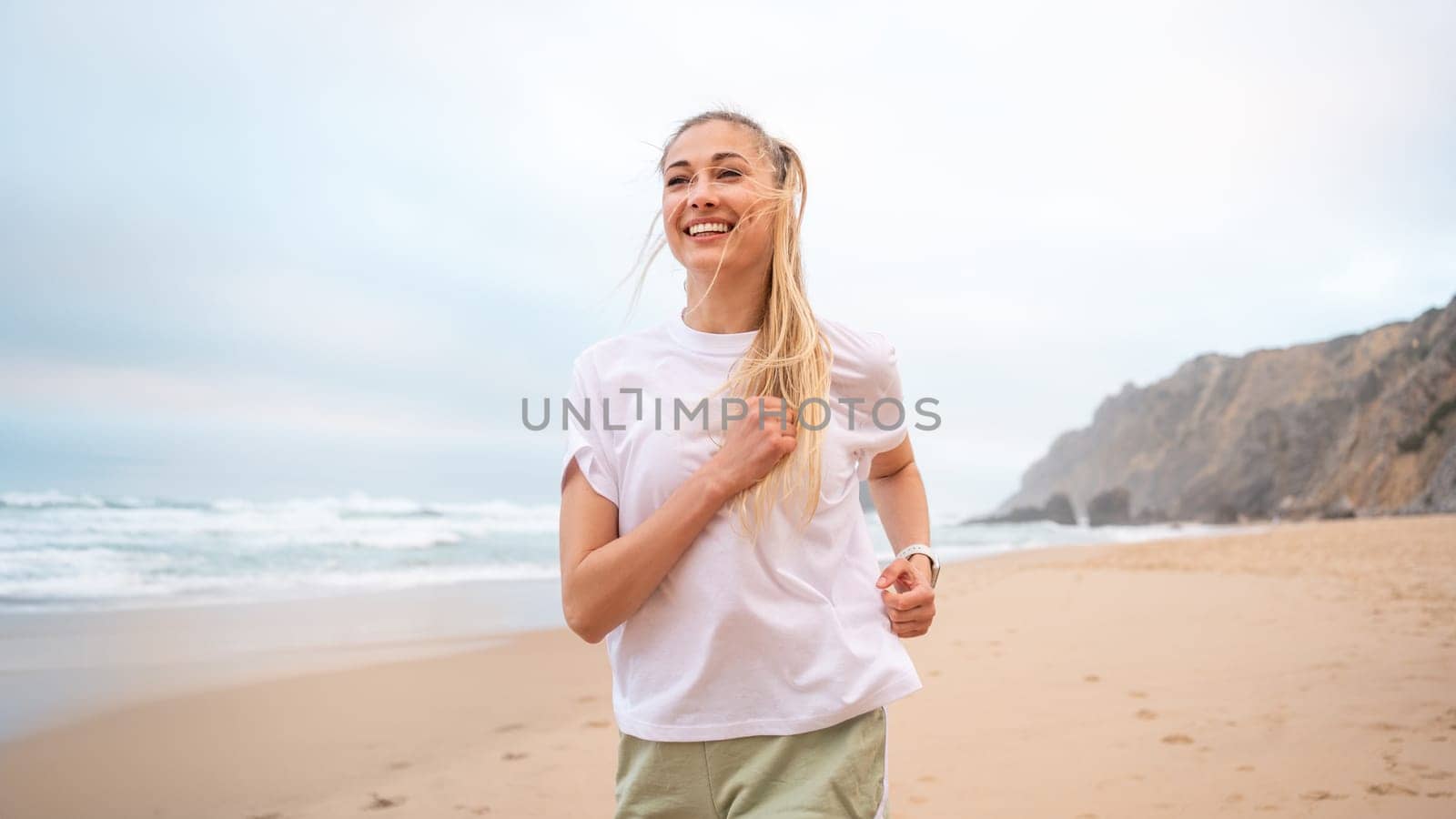 Smiling young woman runner with blond hair in white top jogging on sandy beach. Cheerful female athlete jogging along sea under sky. Happy girl represents healthy lifestyle.