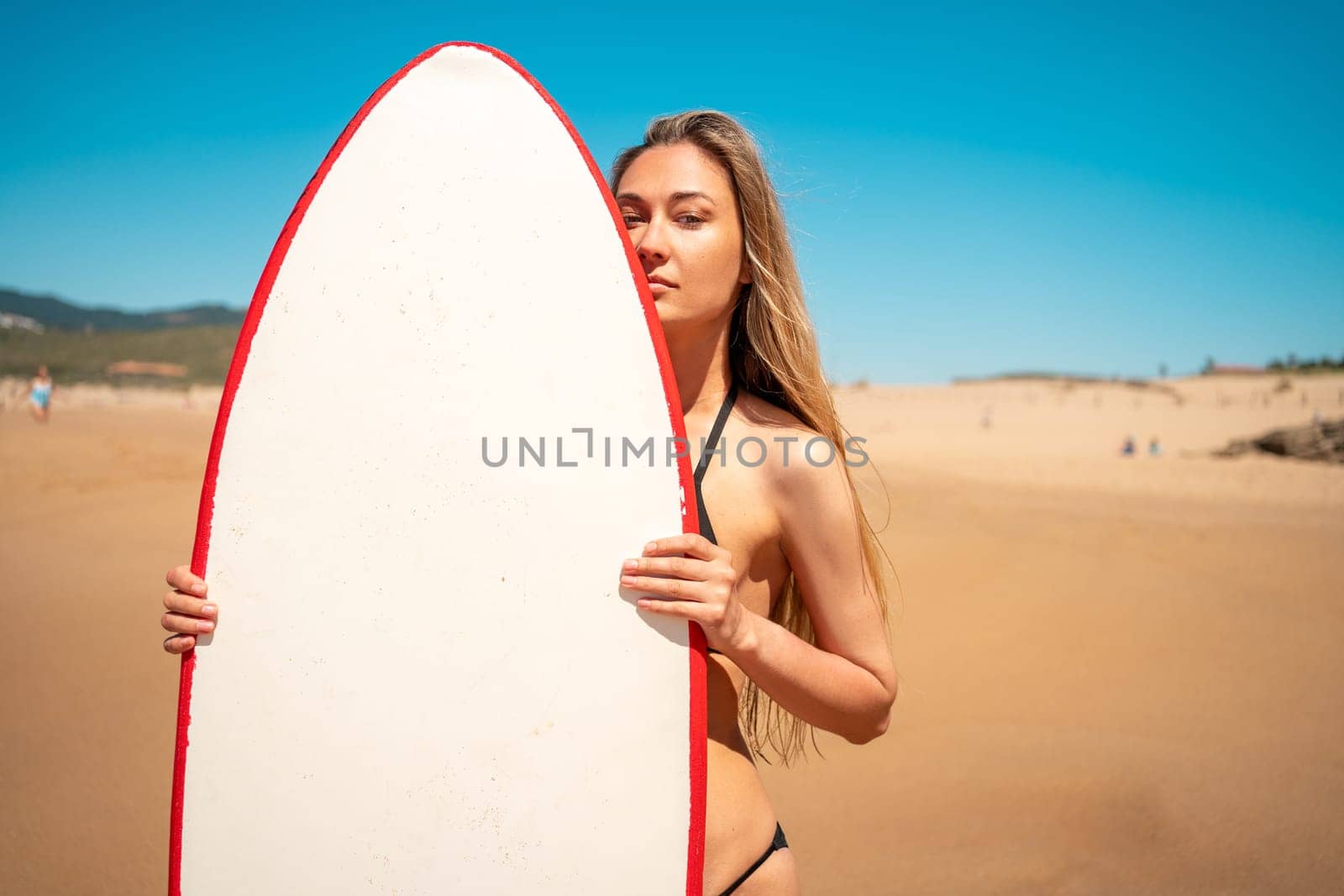 Portrait of attractive young female surfer with surfboard at beach. Confident woman with long blond hair in bikini posing with surfboard near sea under blue sky. Lady is enjoying summer vacation.
