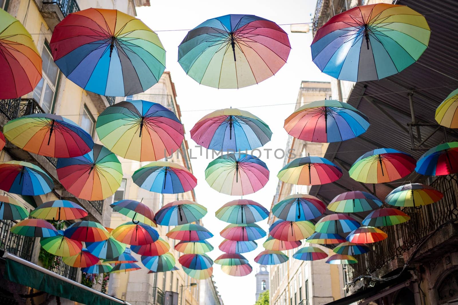 Colorful umbrellas adorn the street, creating vibrant display against blue sky. Many rainbow parasols contribute to festive street decorations, especially during Pride Month.