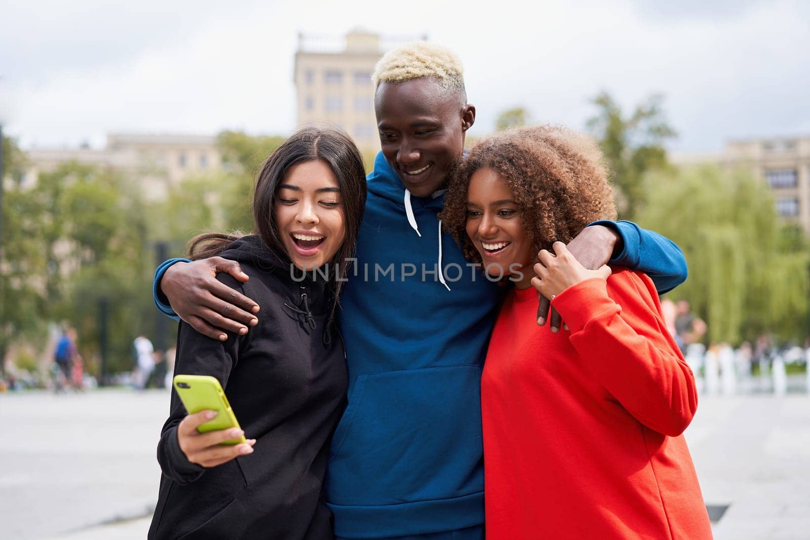 Cheerful diverse young man with female friends standing arm around looking at smartphone screen in city. Happy students of different nationalities spending weekend time together in city park.