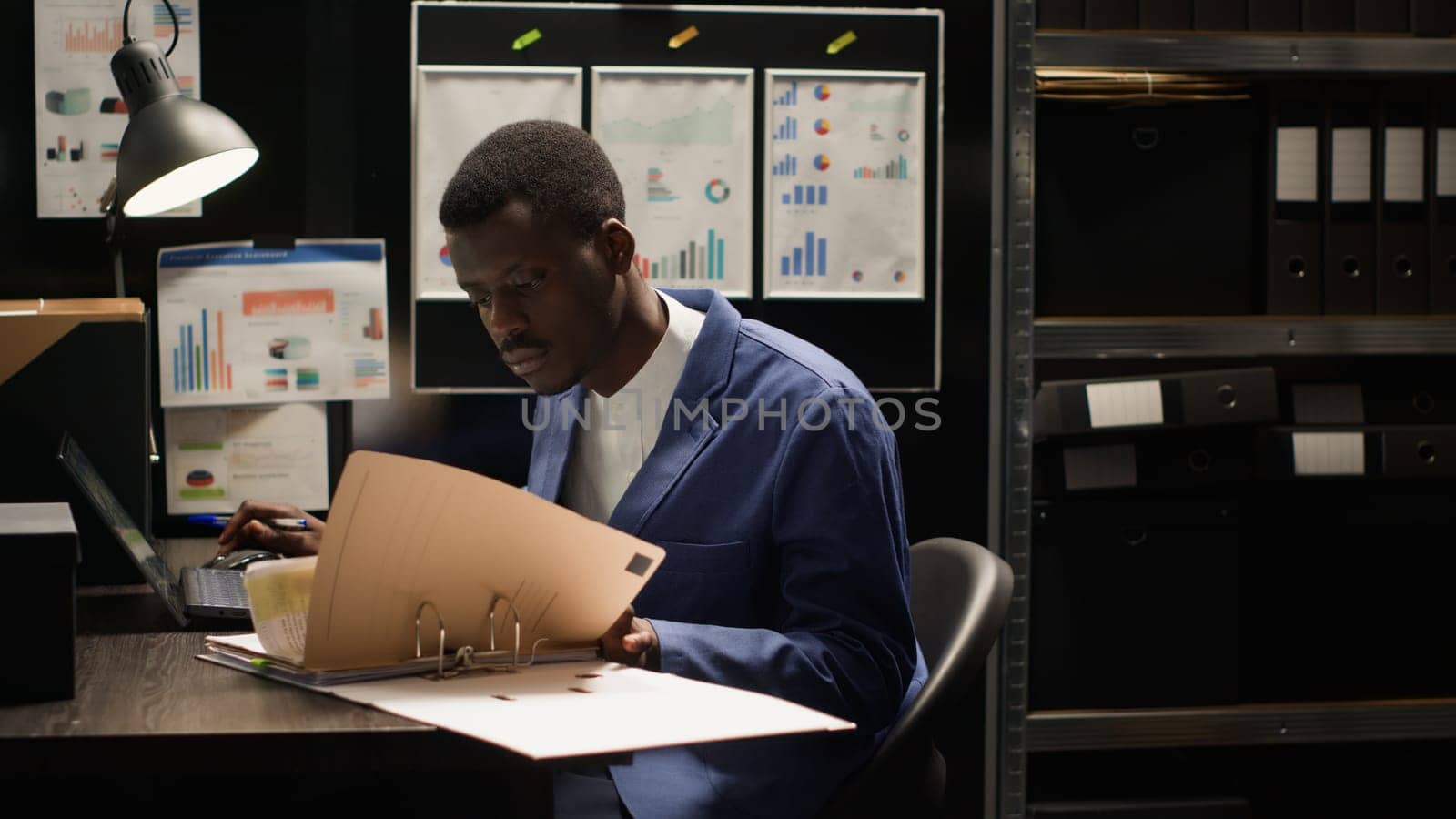 Diligent private investigator grabbing files from shelves to conduct thorough investigation. African american agent with laptop and research papers, analyzes evidence and gathers information.