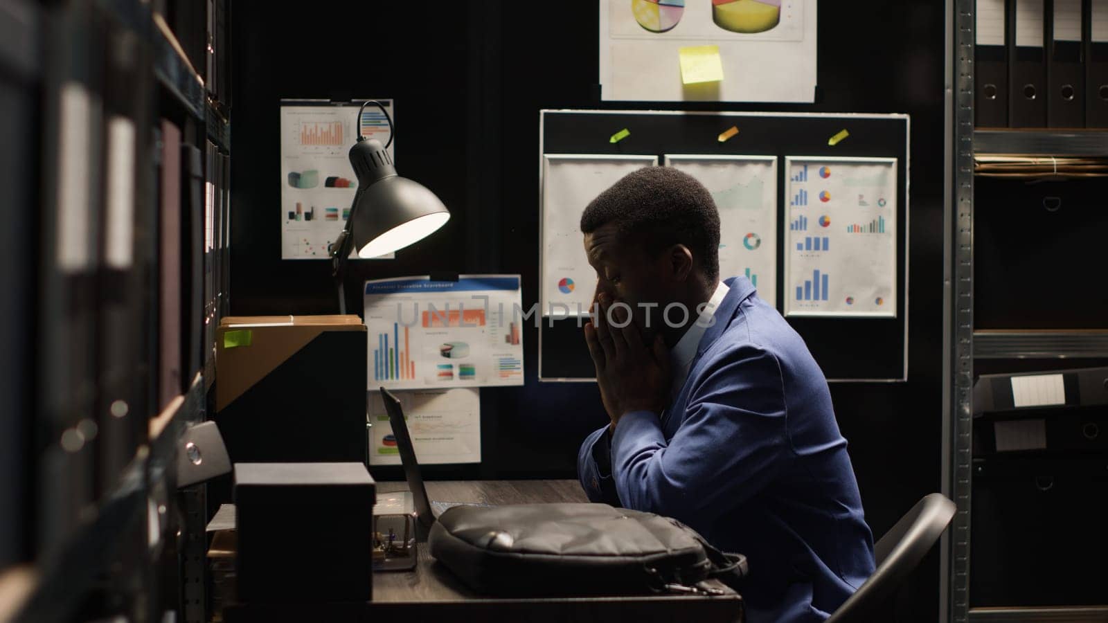 African american law officer with expertise in surveillance, enters discreet office space holding laptop bag. Detective positions laptop on desk and begins reviewing suspect profiles and records.