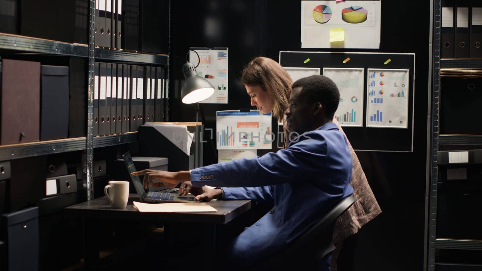 Diverse detectives in well-organized office reviewing evidence, surrounded by shelves filled with files. Two law enforcement officers with laptop, diligently investigate criminal profiles and records.