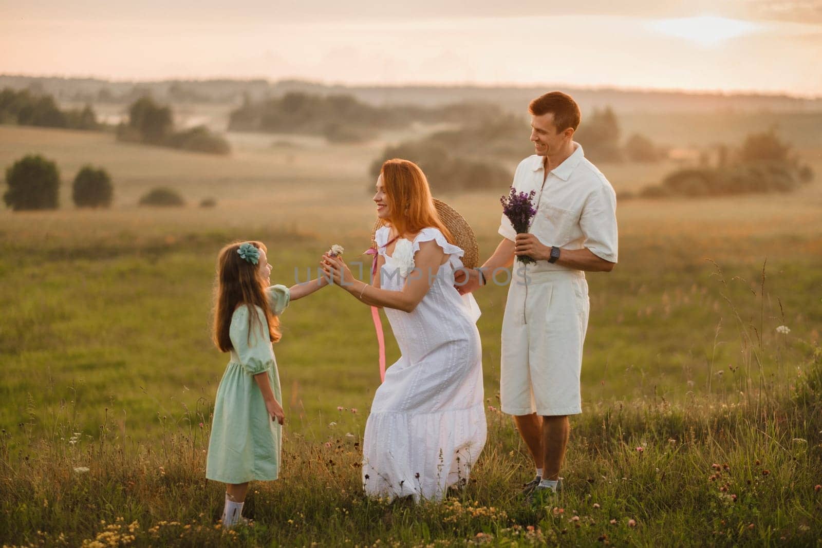 A beautiful happy family of three is standing in a field at sunset.