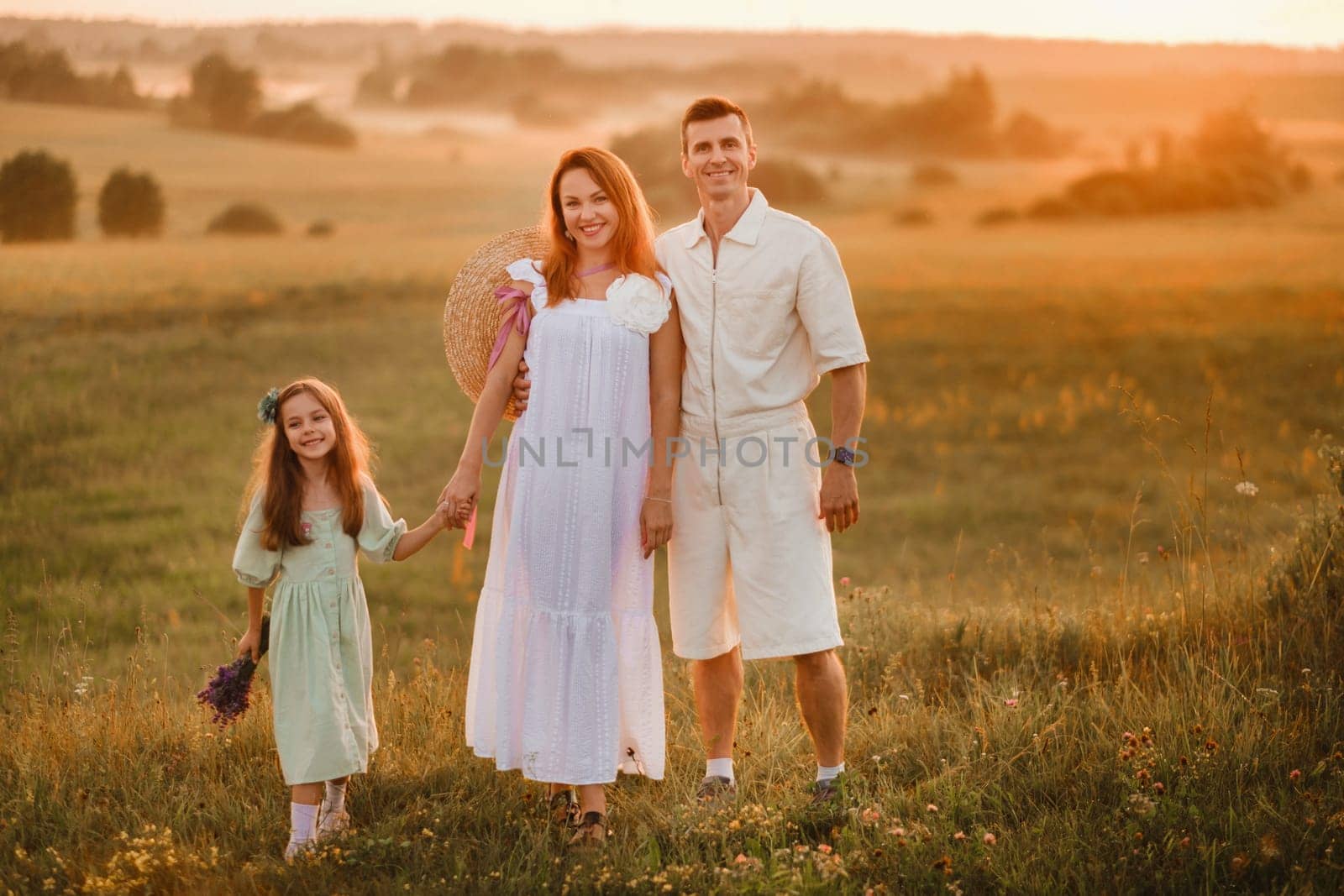 A beautiful happy family of three is standing in a field at sunset by Lobachad