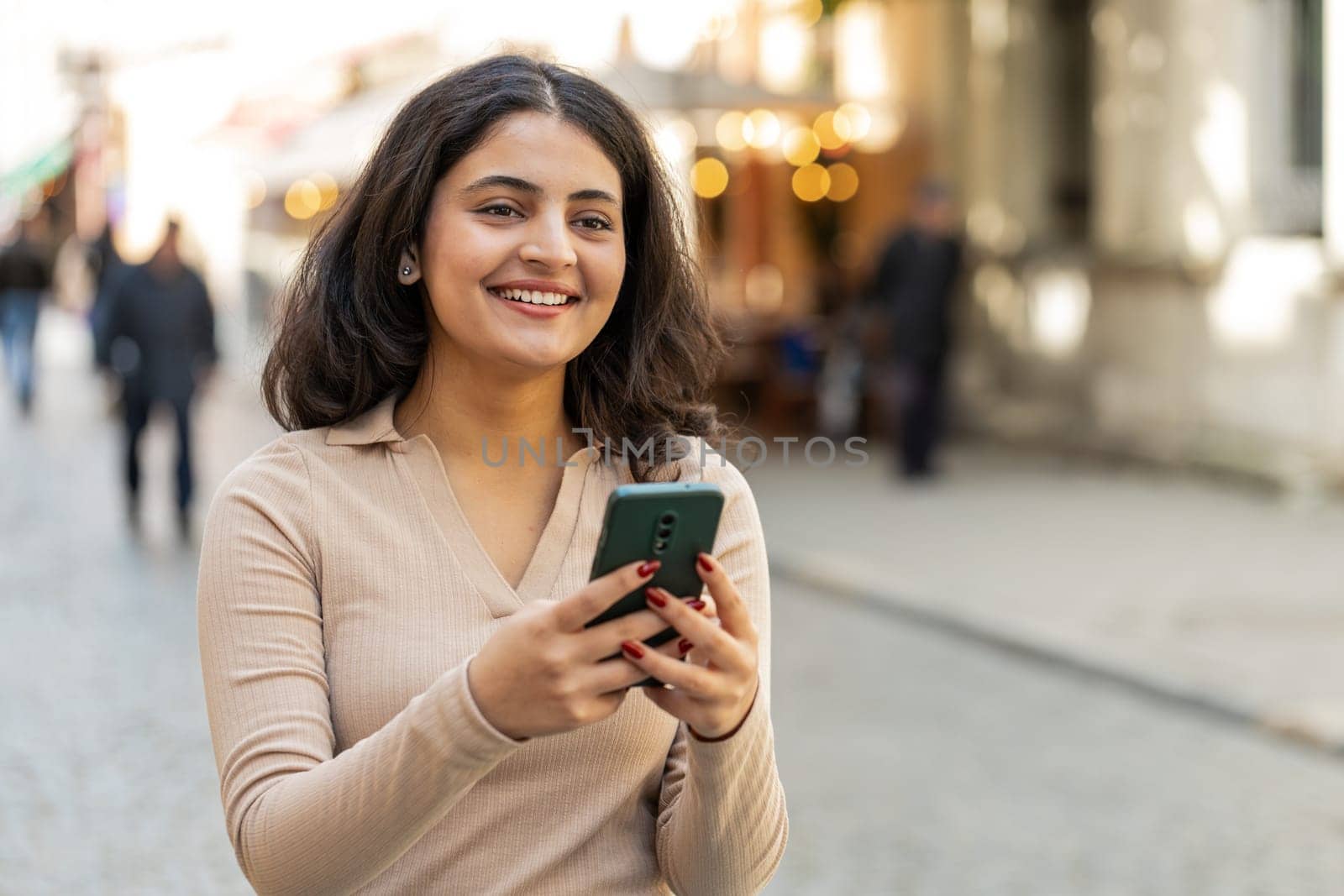 Indian young woman using smartphone typing text answering messages chatting online looking mobile screen social media app outdoors. Hispanic girl holding phone in urban city street. Town lifestyles