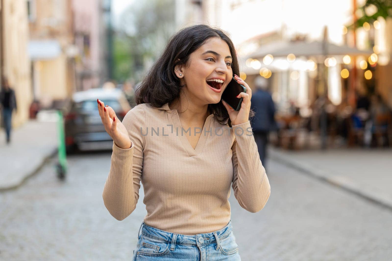 Phone call good news gossip. Happy surprised Indian woman in pleasant conversation on smartphone enjoying talking with friends outdoors. Hispanic girl walking on urban city street. Town lifestyles