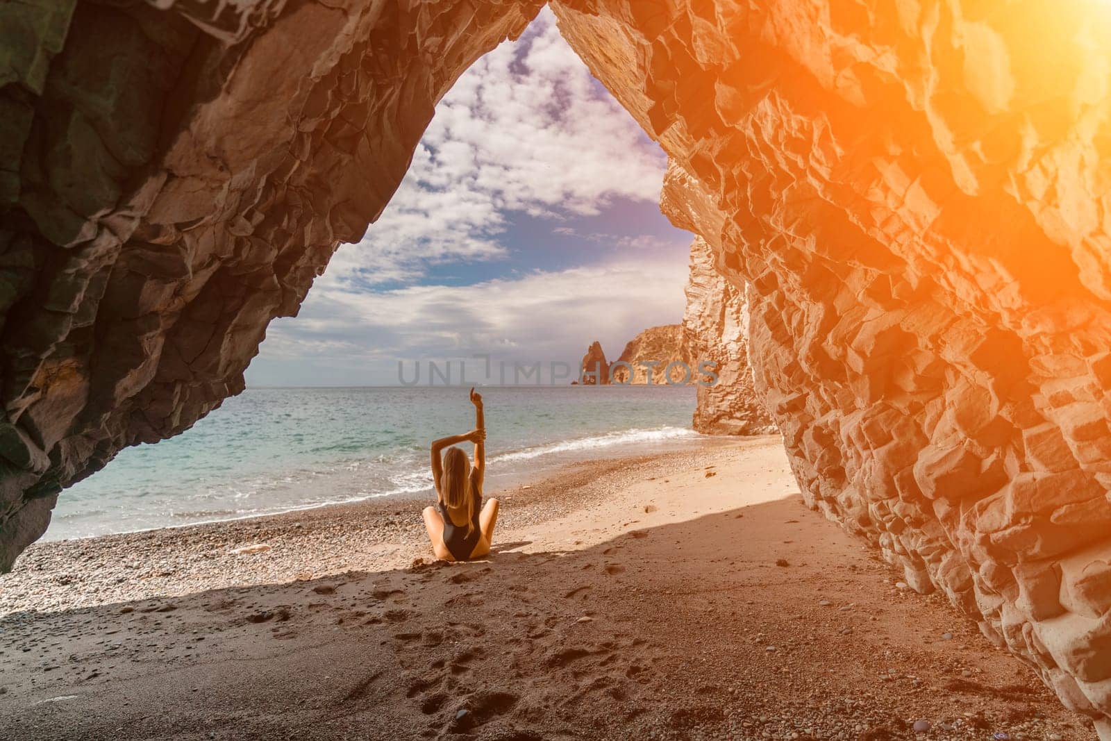 Woman travel sea. View of a woman in a black swimsuit from a sea cave Attractive woman enjoying the sea air sits on the beach and looks at the sea. Behind her are rocks and the sea.