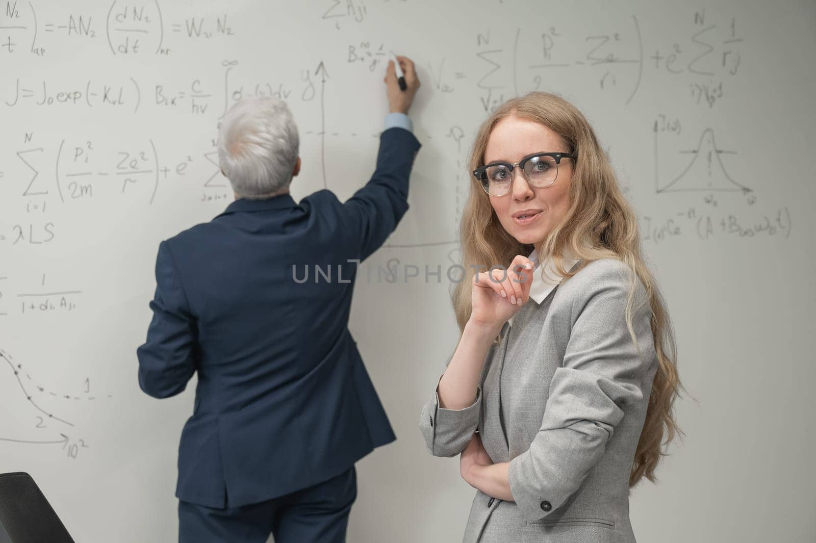 An elderly man writes on a white board and a young woman stands thoughtfully