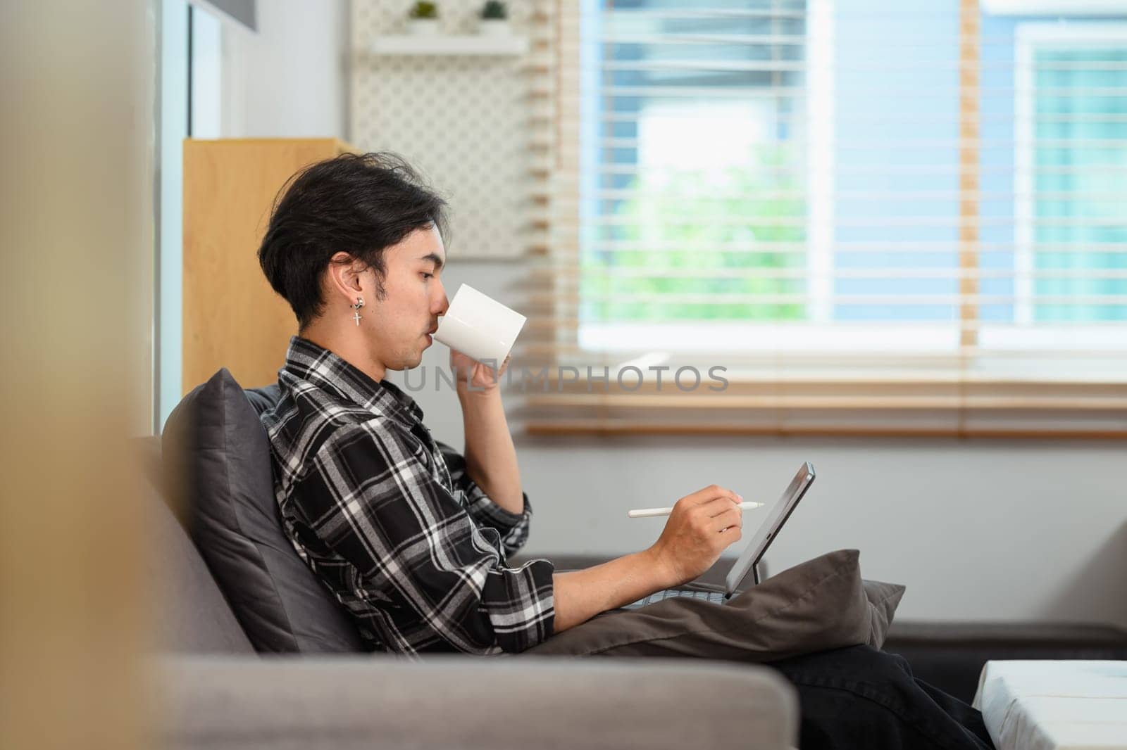 Side view of young man freelancer drinking coffee and using digital tablet in living room.