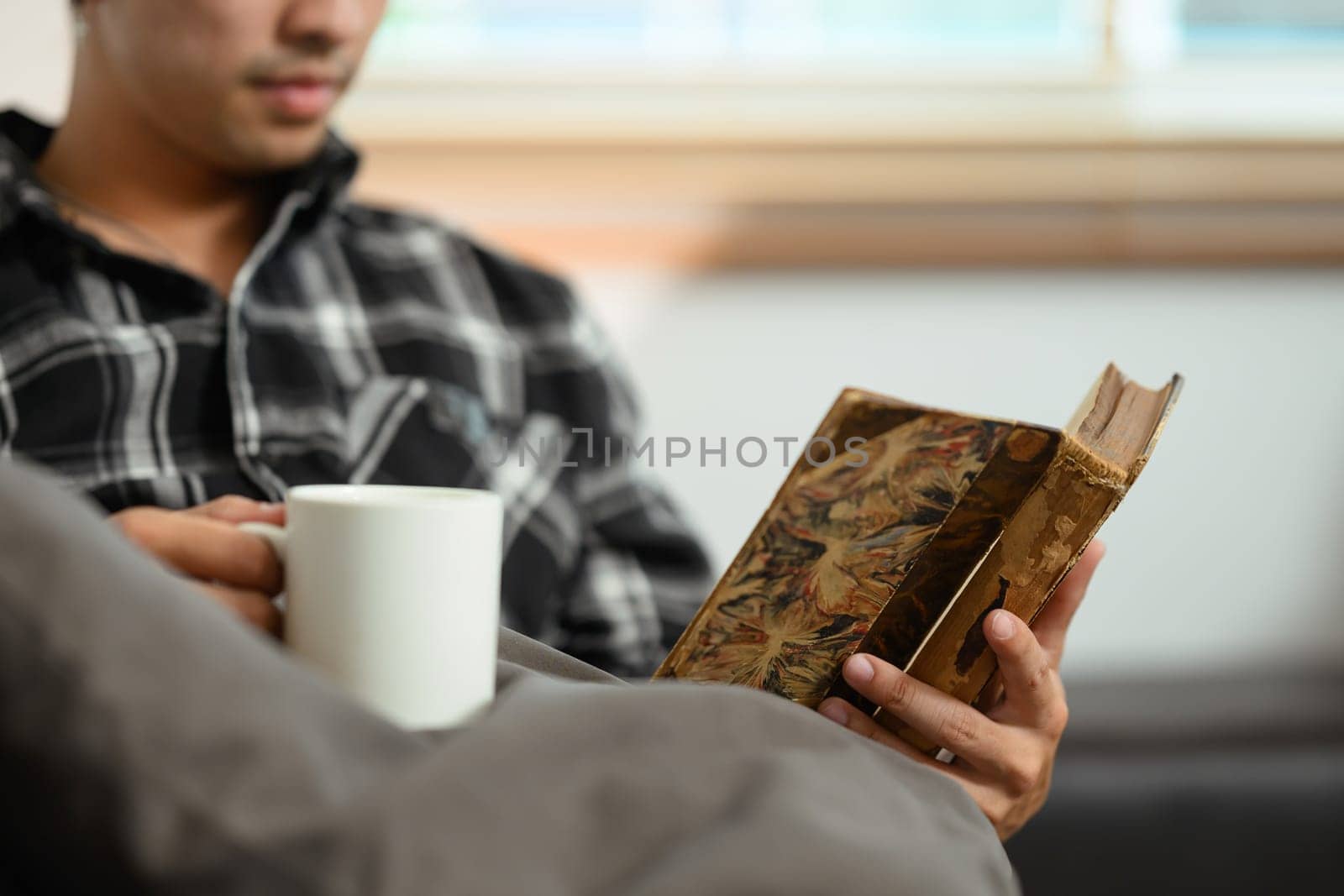 Cropped shot of peaceful young man holding cup of coffee and reading book on couch at home.