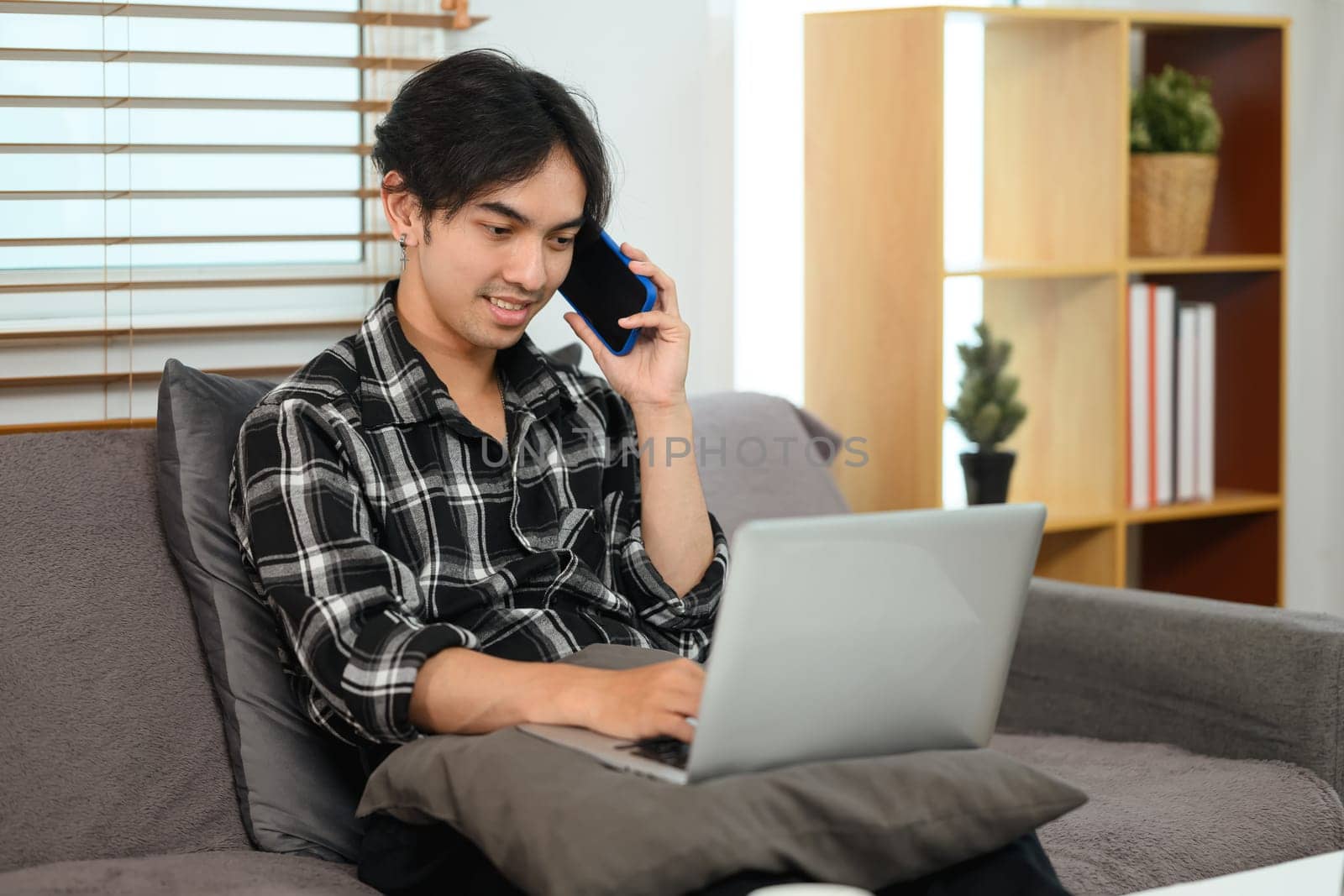 Smiling man working with laptop and talking on mobile phone sitting on couch at home.