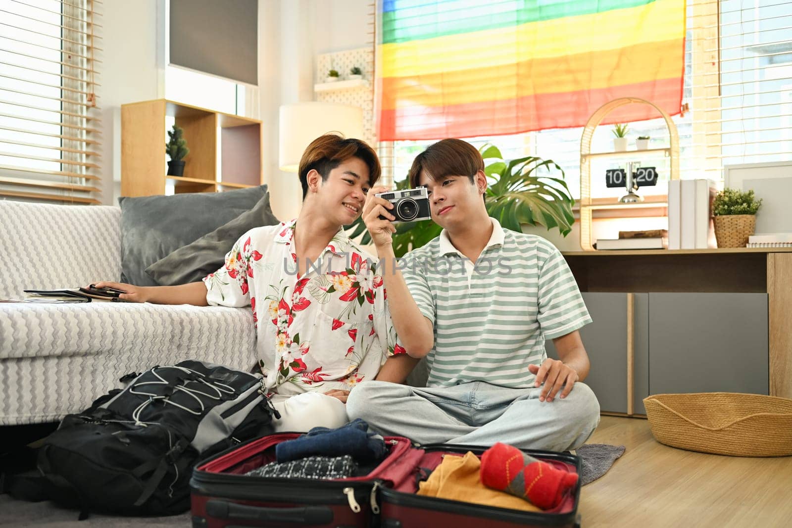 Smiling homosexual couple sitting on floor in living room and packing clothes into travel bag.