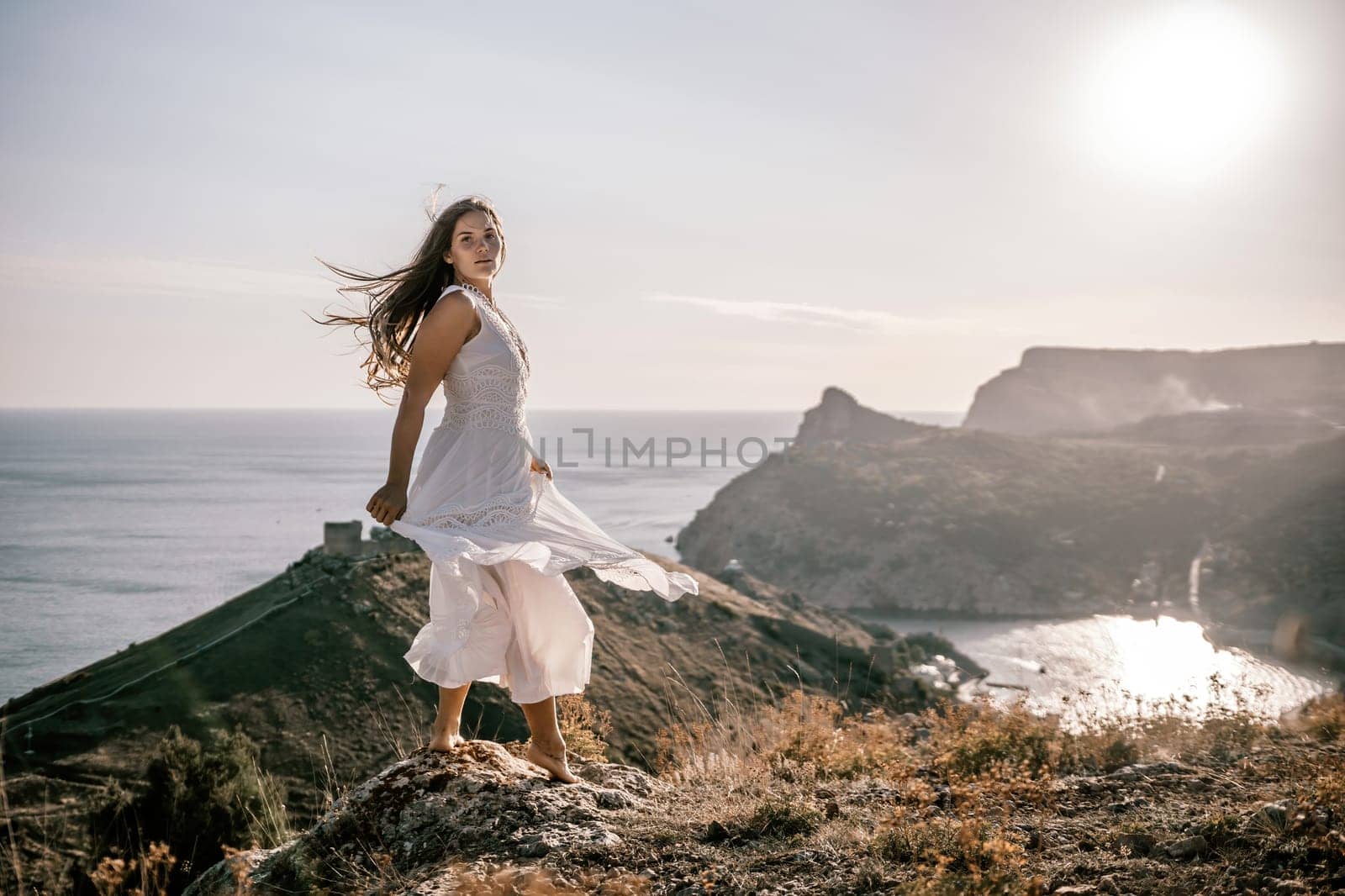 A woman stands on a hill overlooking the ocean. She is wearing a white dress and has long hair. The scene is serene and peaceful, with the sun shining brightly in the background. by Matiunina