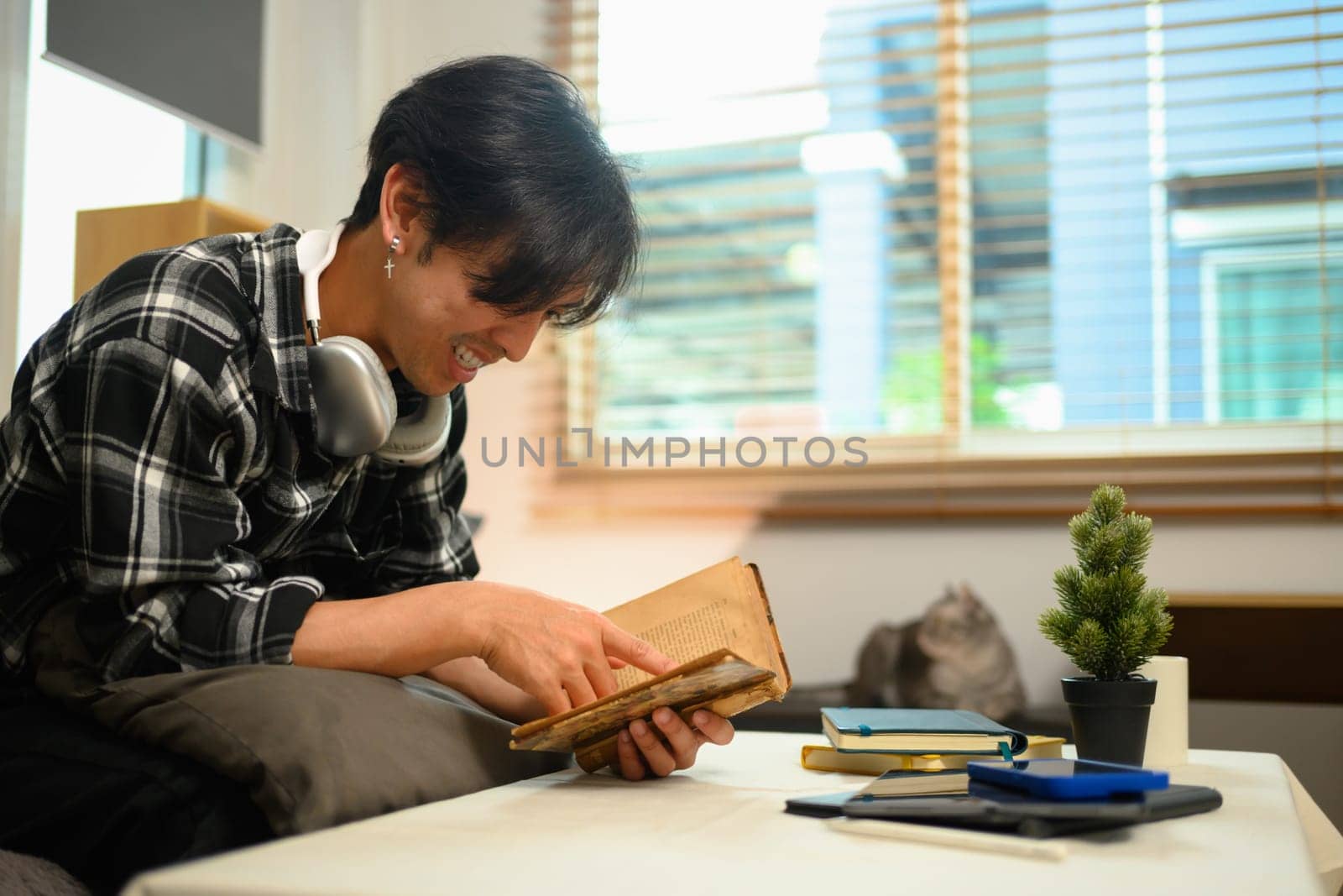 Carefree young Asian man sitting on couch in living room and reading book by prathanchorruangsak