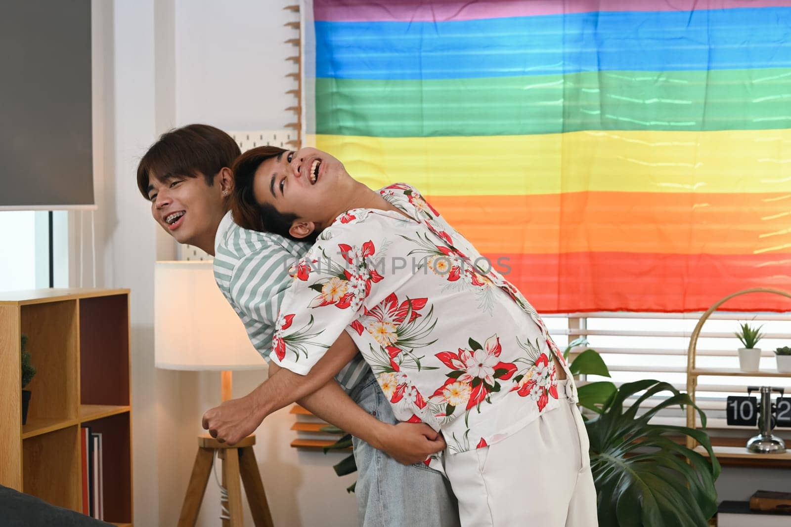Playful gay couple giving piggyback ride for each other laugh joyfully in living room near rainbow LGBT Pride flag.