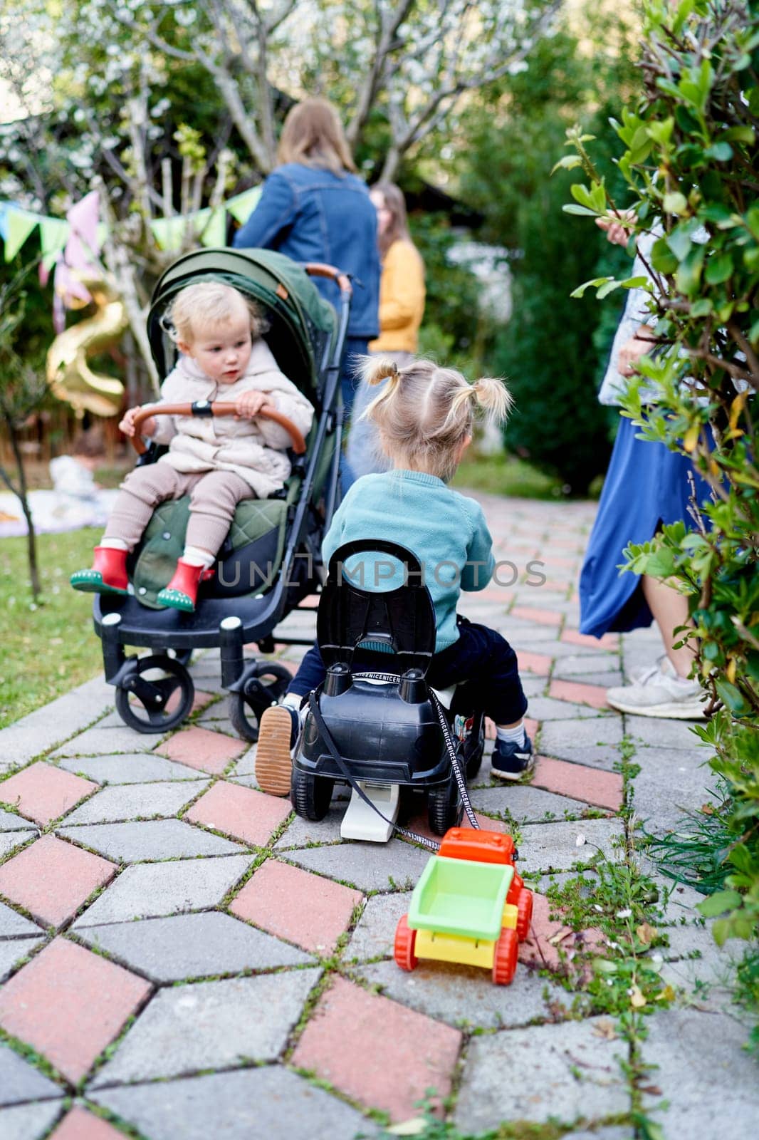 Small child sits in a stroller and looks at a little girl driving a toy car with a small truck tied to the seat. High quality photo