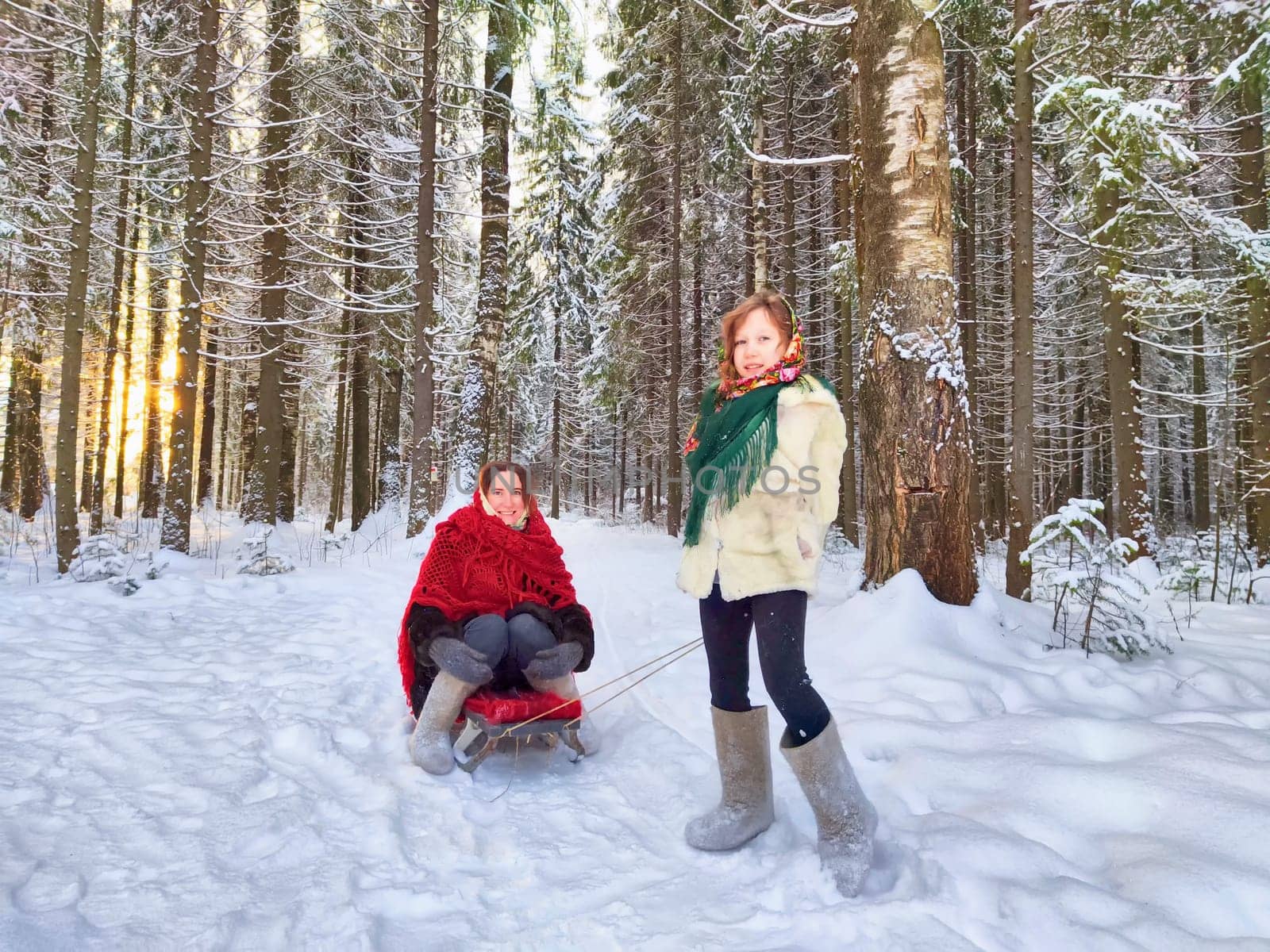 A mother and daughter in shawls are sledding in the winter forest. Ethnic clothes and fun on Shrovetide carnival Maslenitsa by keleny