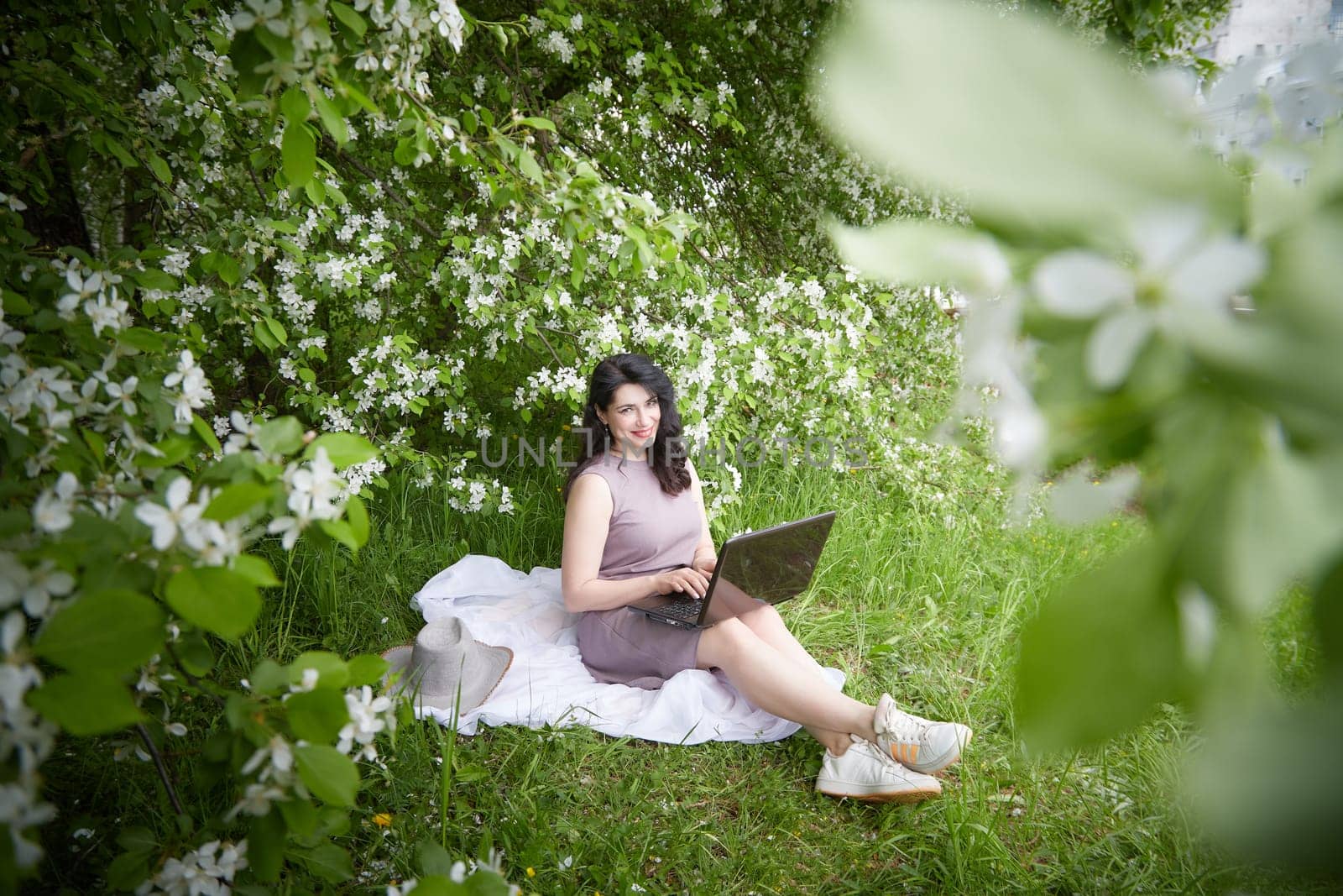 Girl engages with laptop amidst lush greenery. Freelancer Woman Working on Laptop Outdoors in a Blooming Garden
