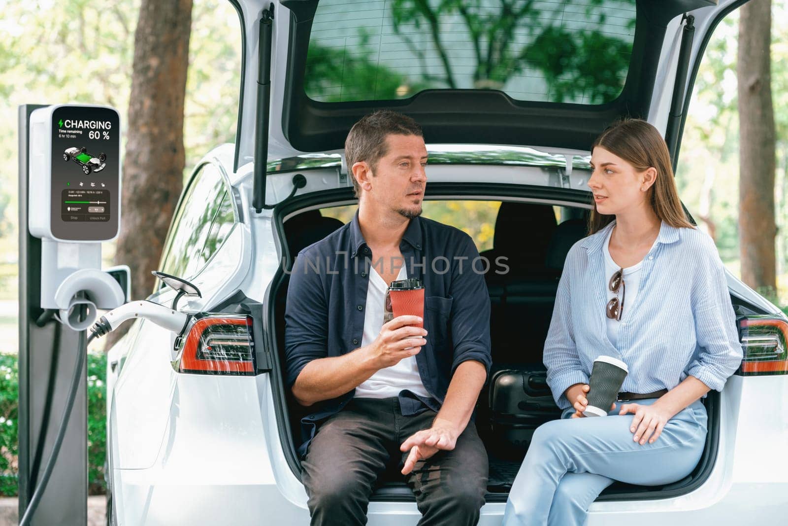 Lovely young couple drinking coffee while recharging battery for electric car during road trip travel EV car in natural forest or national park. Eco friendly travel during vacation and holiday. Exalt