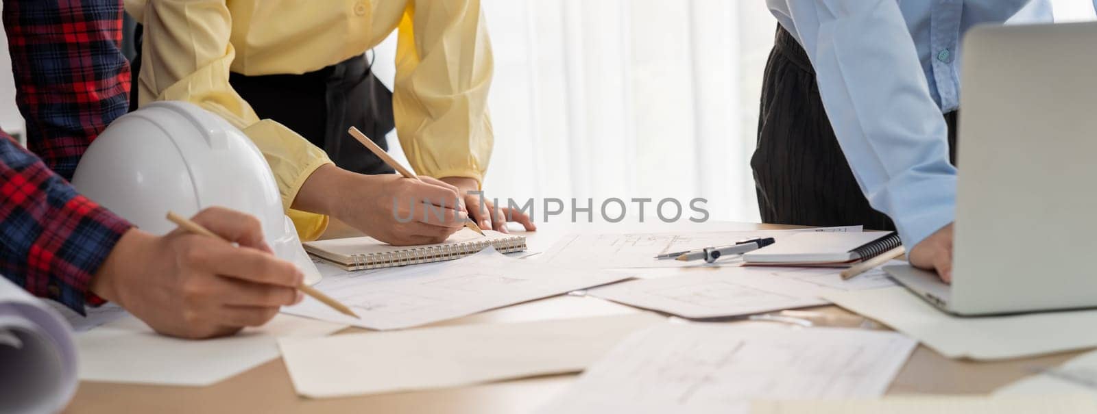 Professional engineer draws a blueprint on meeting table with blueprint and equipment scattered around while project manager using laptop analysis data at meeting room. Closeup. Delineation.