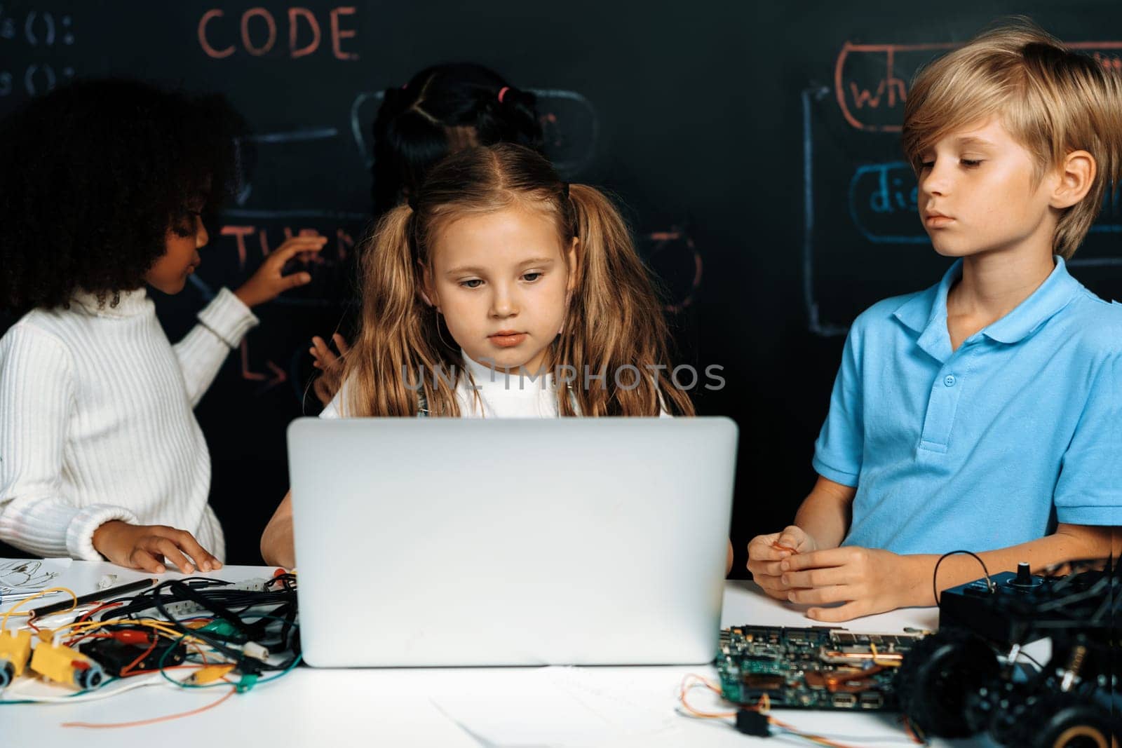 Smart girl in white bib learning about coding robotics technology using laptop in the STEM class. Schoolboy in blue shirt try to educate motherboard while smart schoolgirls reading code. Erudition.
