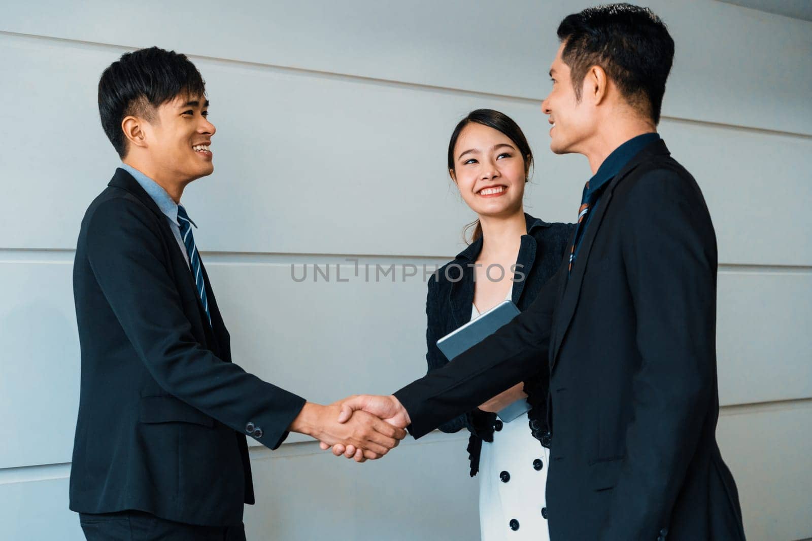 Business people agreement concept. Businessman do handshake with another businessman in the office meeting room. Young Asian secretary lady stands beside them. uds