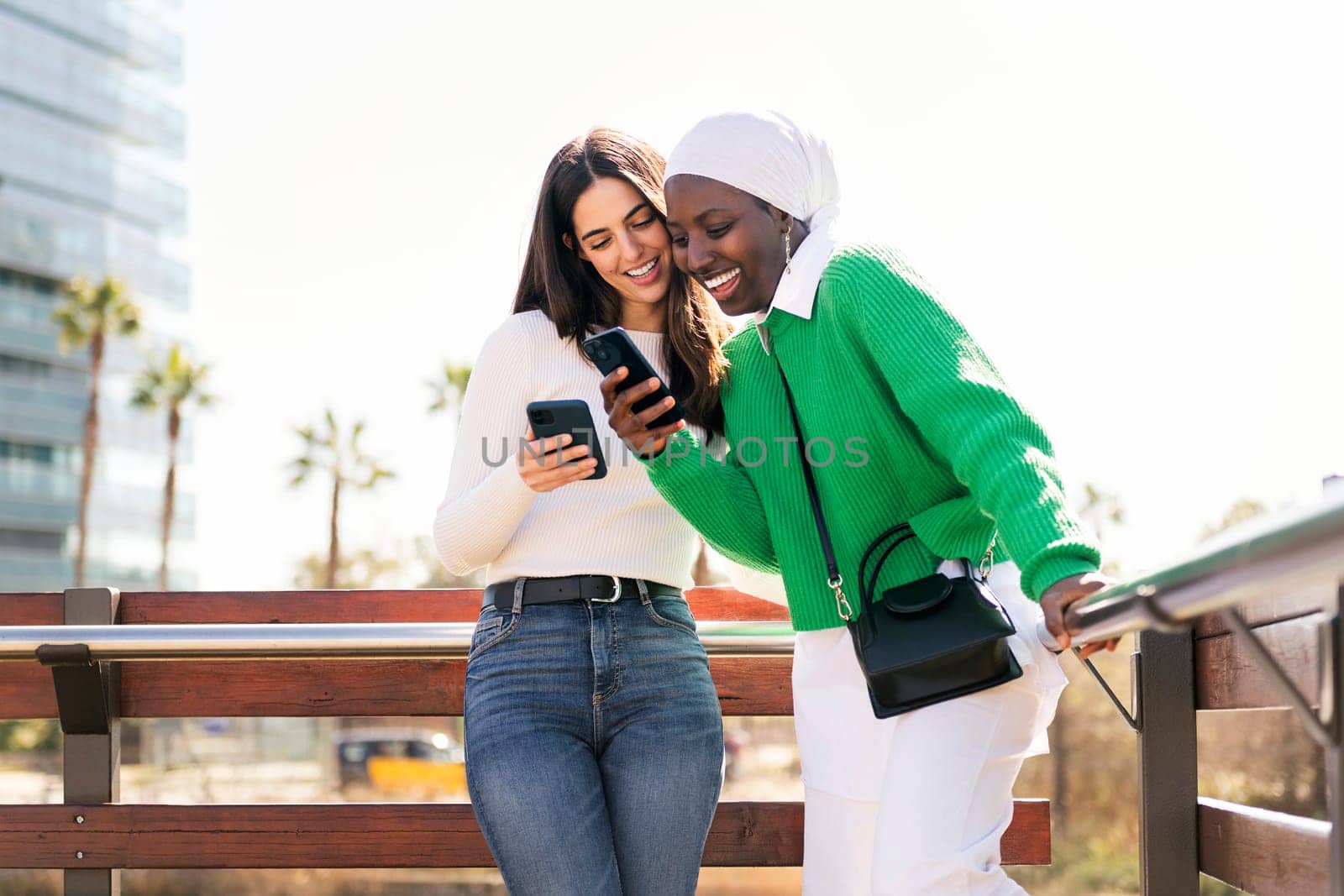 two young female friends smiling happy while having fun carefree looking at their mobile phones in a city park, concept of diversity and modern lifestyle, copy space for text