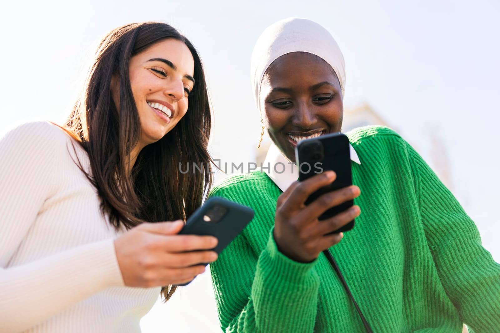 two young female friends smiling happy while having fun carefree looking at their mobile phones, concept of diversity and modern lifestyle