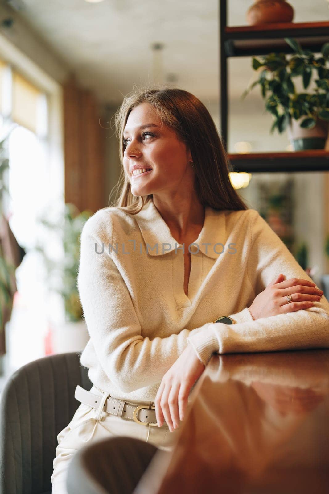 A young woman with a subtle smile is standing in a warmly lit cafe. She exudes confidence and a casual elegance, dressed in a chic white blouse. The cafes casual atmosphere is enhanced by potted plants and natural light.