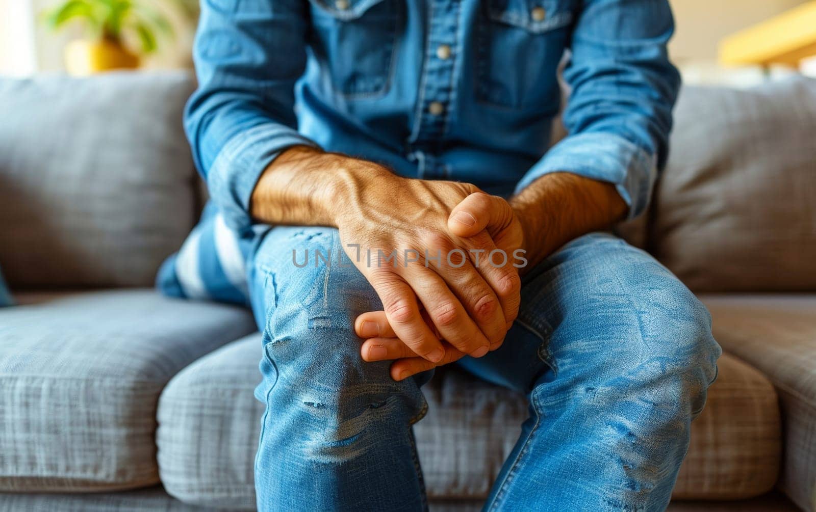 A man sits casually on a couch, hands clasped firmly over his knees, wearing a denim outfit. His focus is drawn to his hands, reflecting a moment of deep thought or concern.. by sfinks