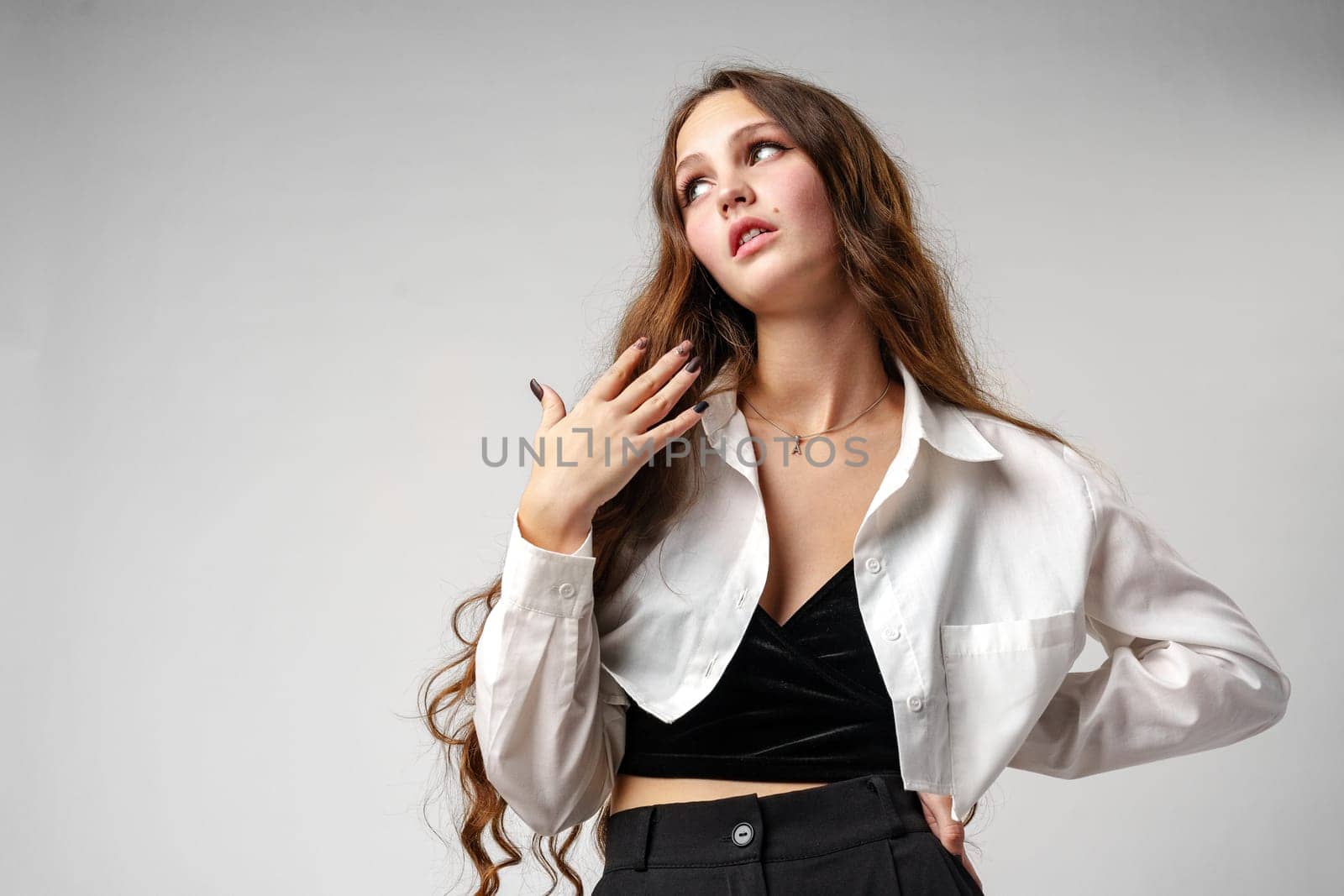 A woman with long, wavy hair stands confidently against a neutral backdrop, her head tilted upward in a pose of contemplation or aspiration. She is wearing a fashionable white oversized shirt with the collars slightly raised, layering over a sleek black top which contrasts with her pale skin. Her hand lightly touches her collarbone, adding an elegant touch to her overall poised demeanor.