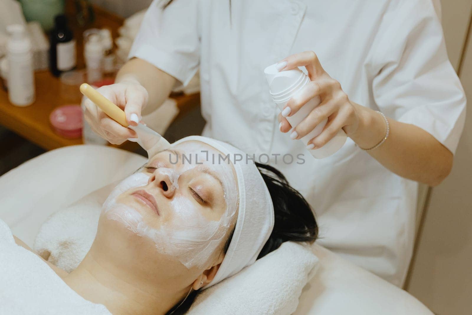 A girl cosmetologist applies a cream mask with a brush to a woman s face. by Nataliya