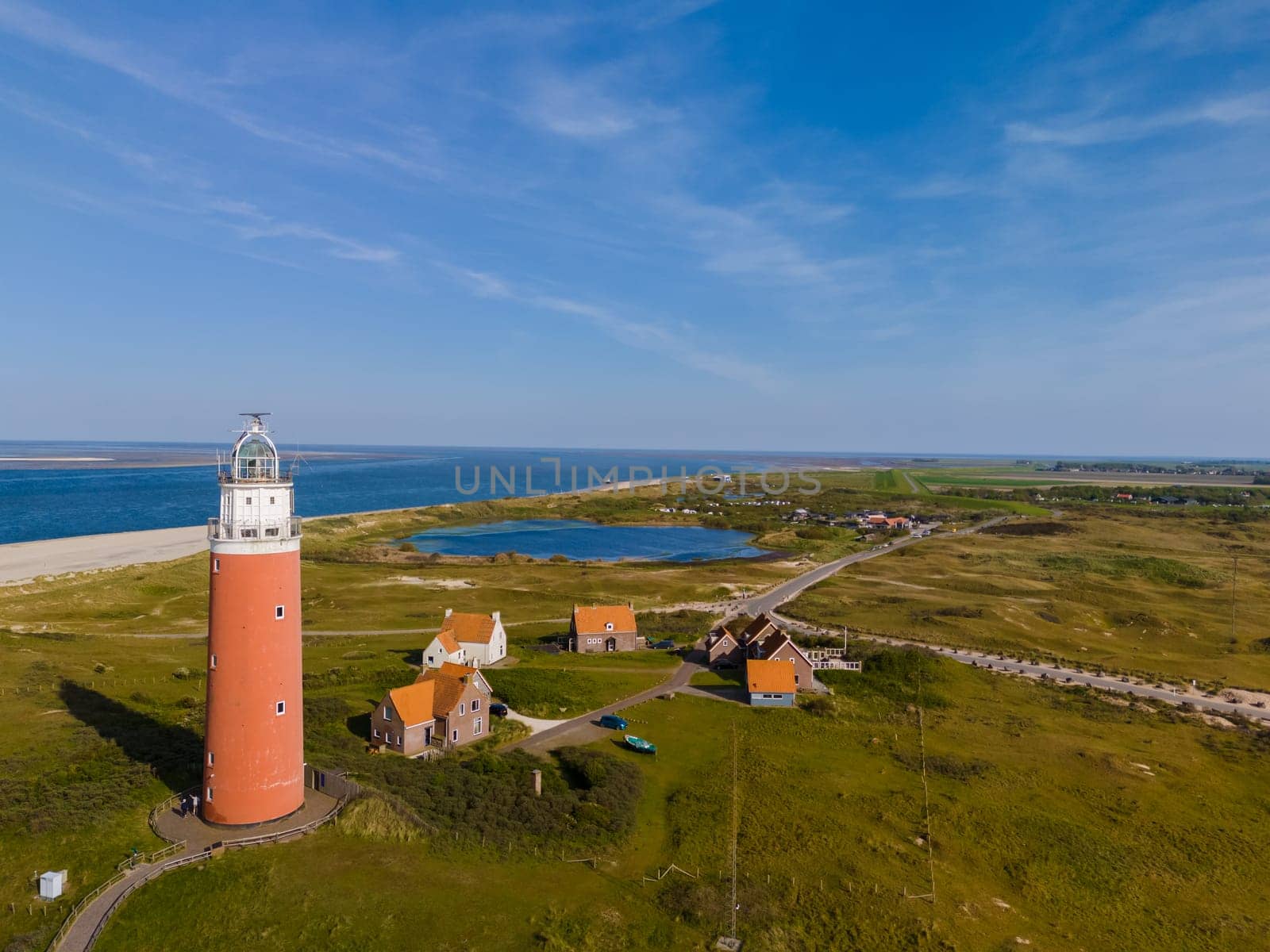 An aerial perspective showcases a majestic lighthouse standing tall near the sandy shores, overlooking the vast ocean. The iconic red lighthouse of Texel Netherlands