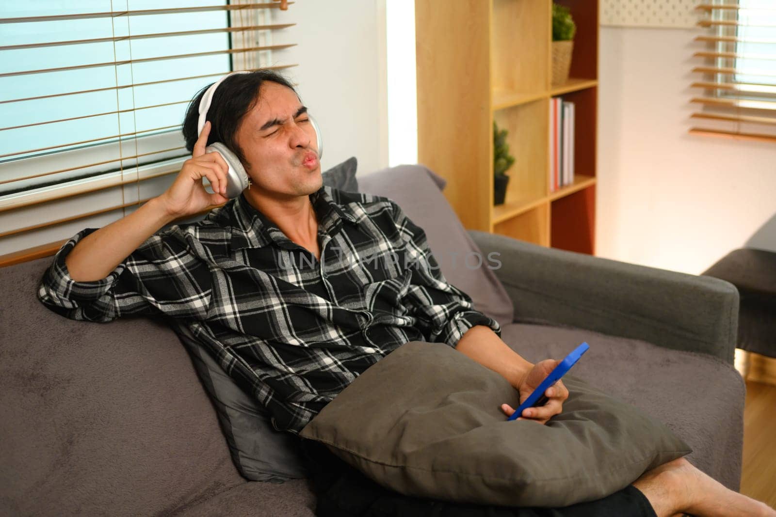 Cheerful asian guy holding mobile phone enjoying favorite songs while relaxing on sofa at home.