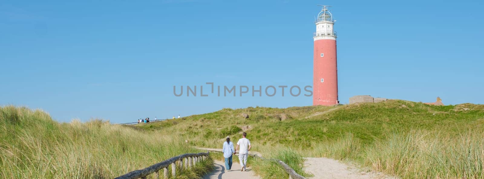 Two people walk down a path next to the lighthouse in Texel, Netherlands, with waves crashing against the shore in the background. a couple at The iconic red lighthouse of Texel Netherlands